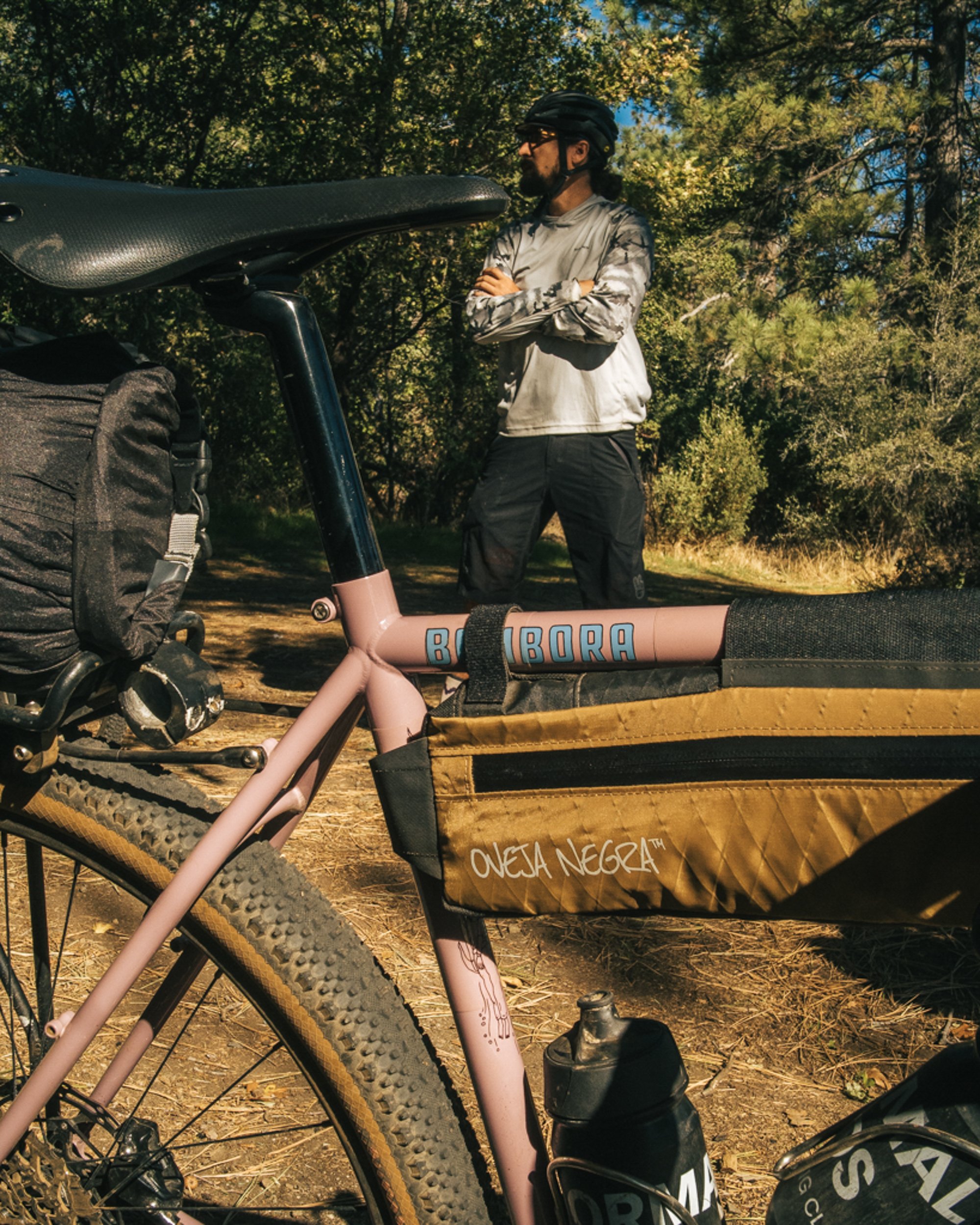 A person standing with arms crossed, wearing a helmet and sunglasses, is outdoors near a pink bike with a black and yellow storage bag attached. The background consists of trees and foliage.