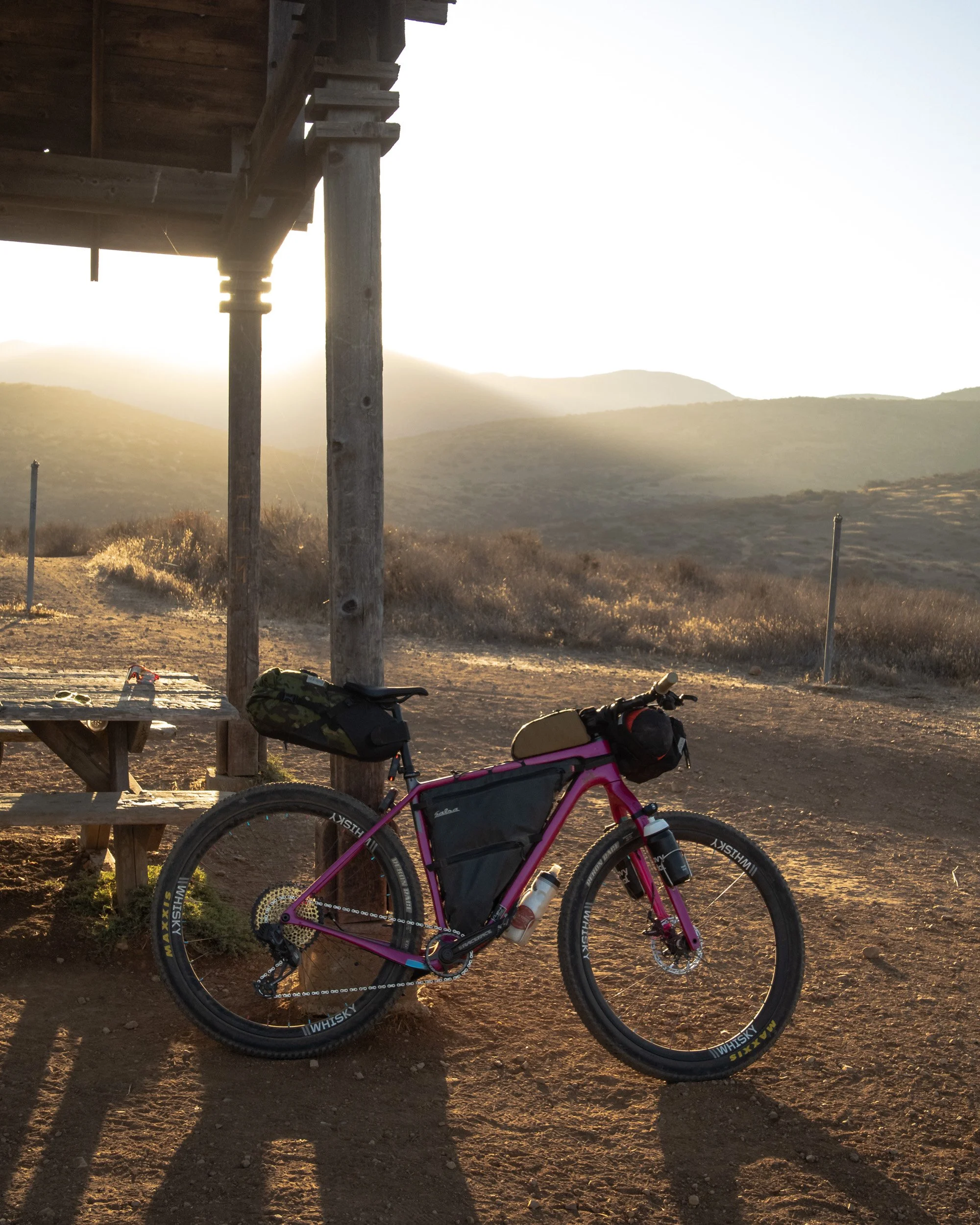 A pink mountain bike with gear bags parked near a wooden shelter on a dirt trail, with hills and a sunset in the background.