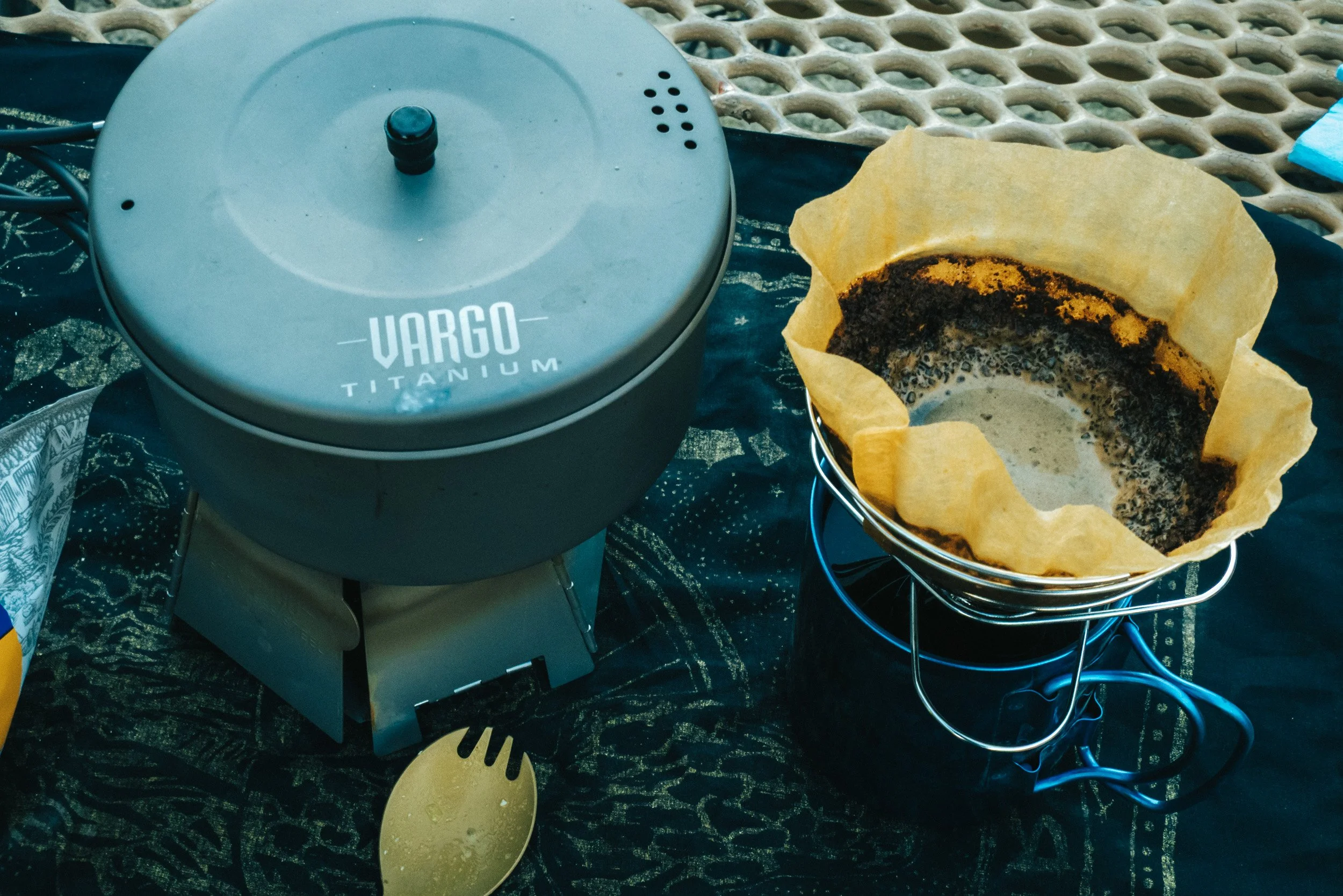 A portable coffee roaster labeled Vargo Titanium next to a coffee filter with used coffee grounds on a metal stand, placed on a dark cloth surface outdoors.