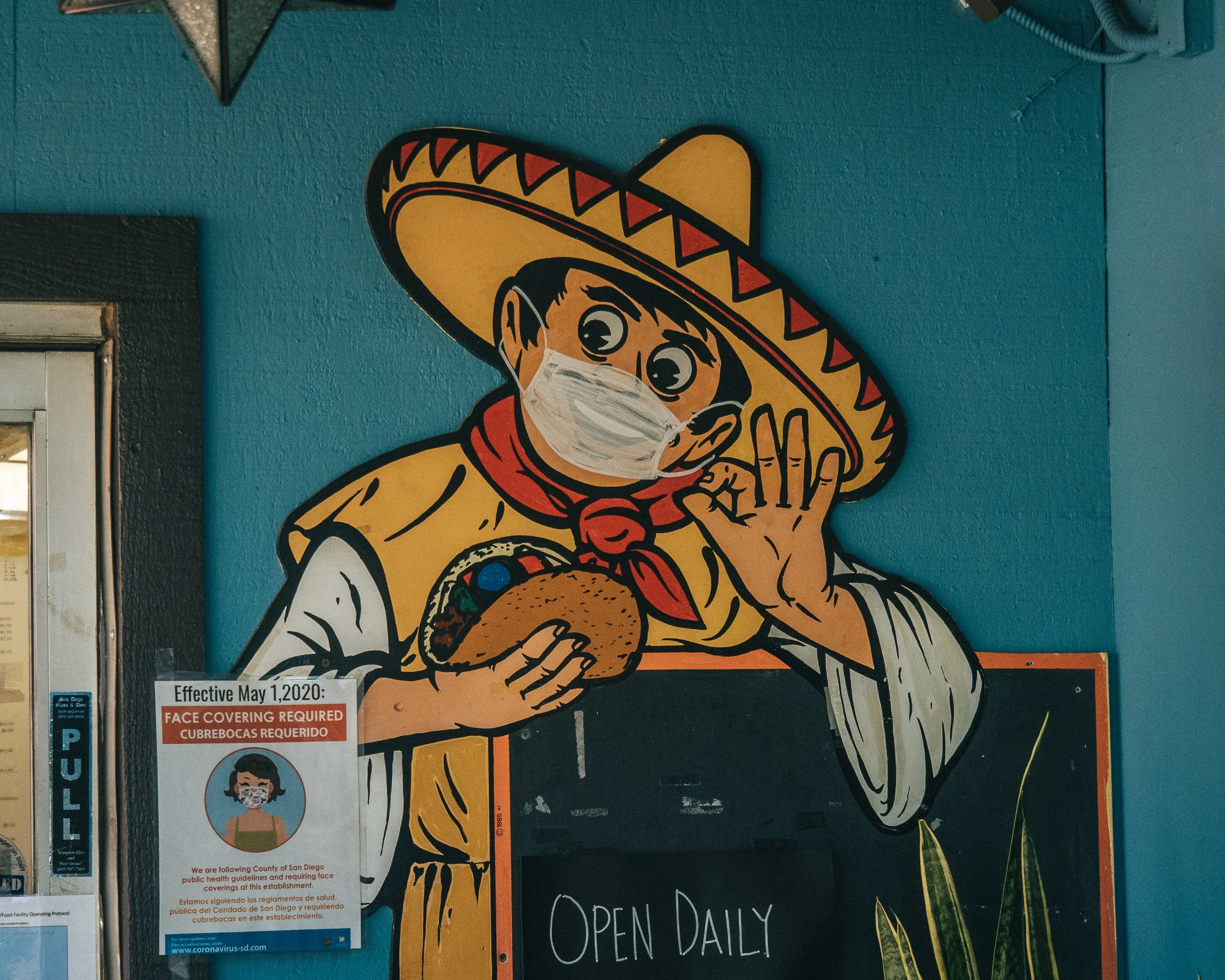 Cartoon of a person wearing a large sombrero, a yellow poncho, holding a taco, and wearing a face mask, making an okay hand gesture.