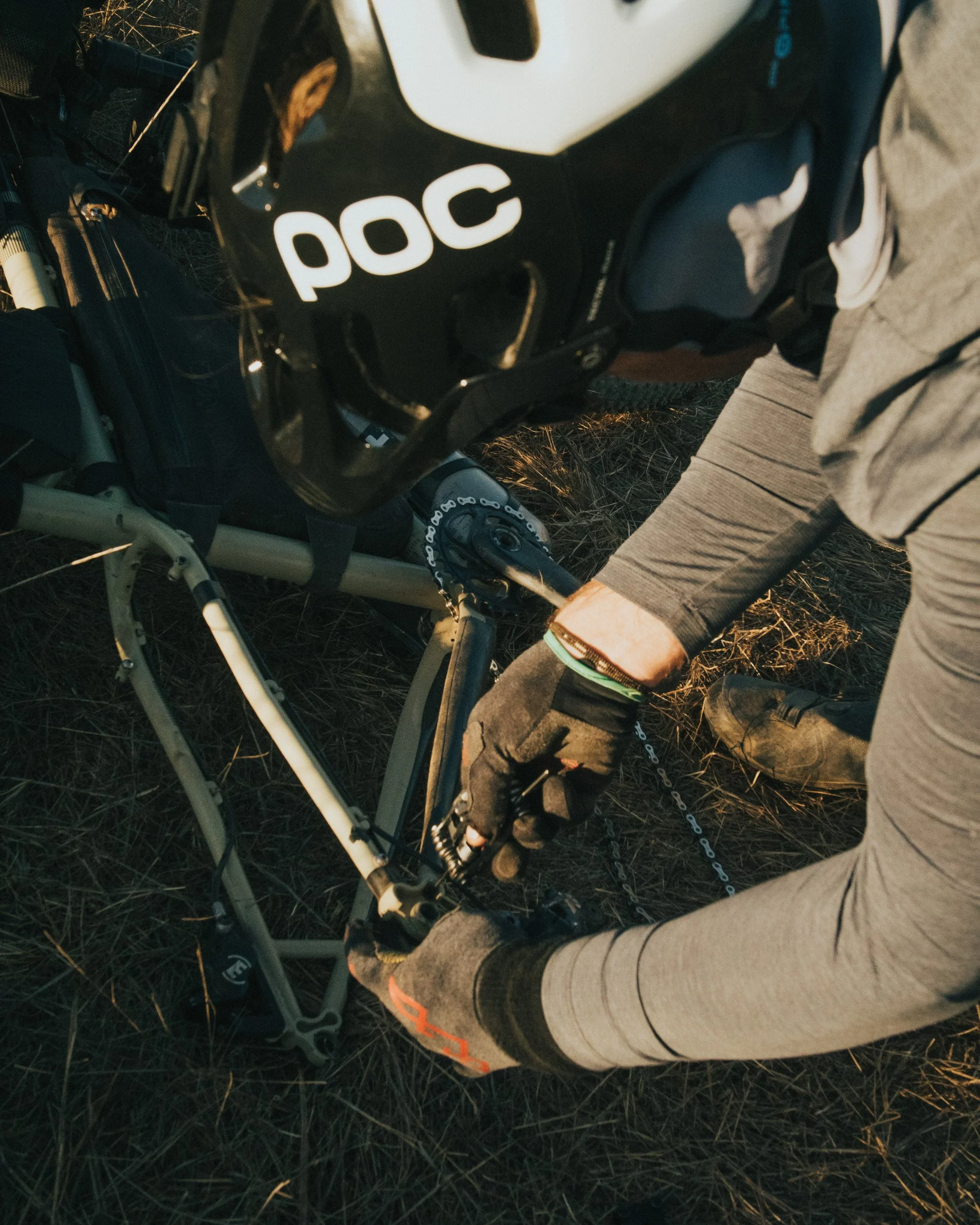 A person wearing a helmet with 'POC' branding and gloves is repairing or adjusting a mountain bike on a grassy field.