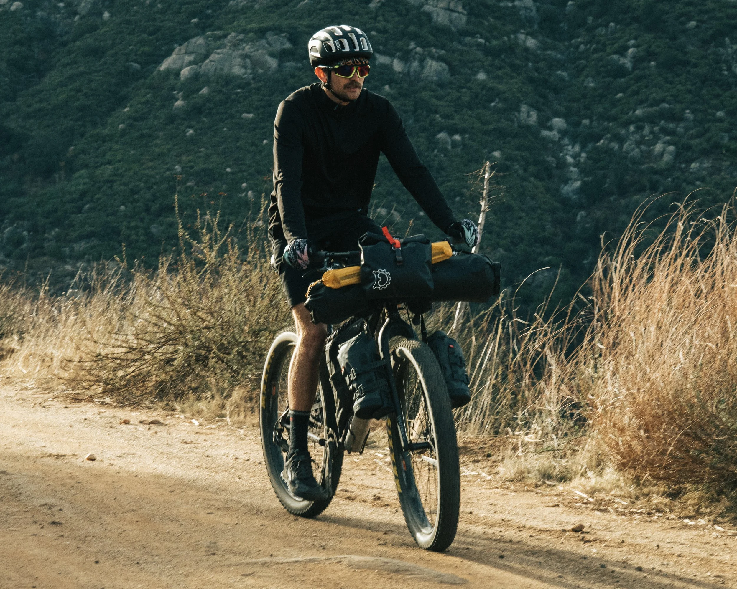 A man riding a mountain bike on a dirt trail in a dry, grassy area with hills in the background.
