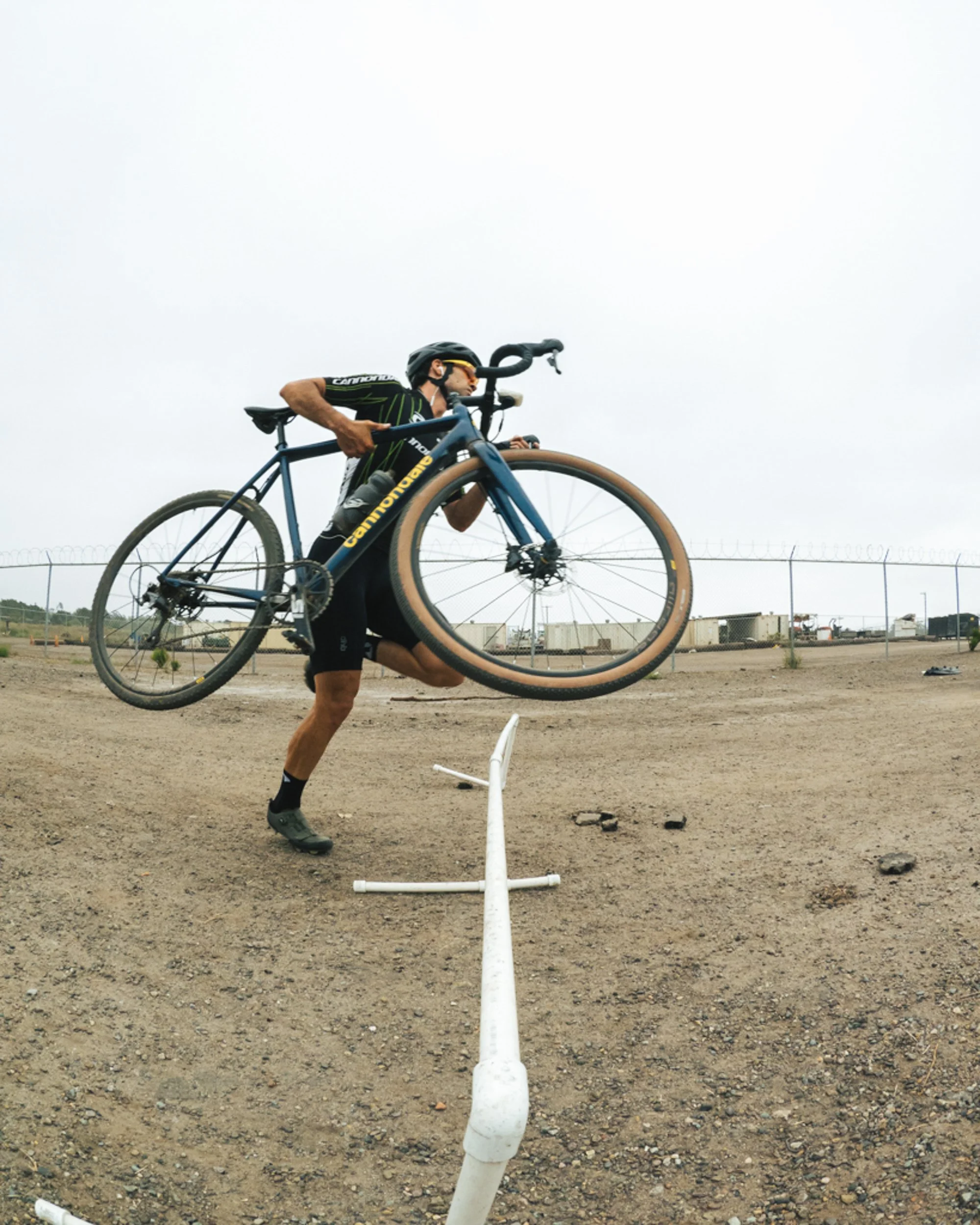 A cyclist is jumping over a white barrier while holding a blue Cannondale road bike in mid-air, wearing a black helmet, black cycling gear, and sunglasses, on a dirt field with a fence and some trailers in the background.