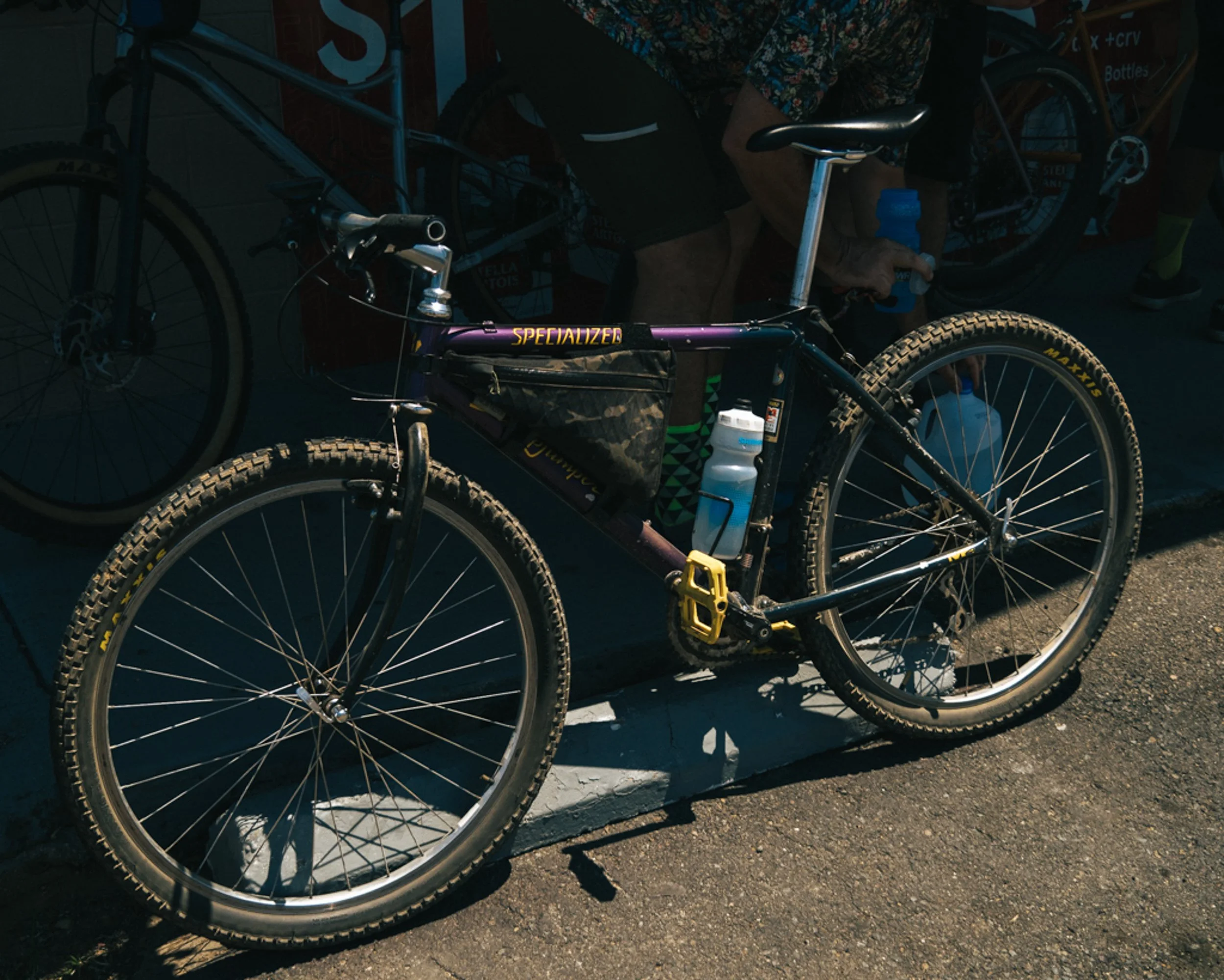 Close-up of a purple Specialized mountain bike with a water bottle attached, parked on the street with a shadow cast over it. In the background, a person with a bicycle is bending over, partially visible.