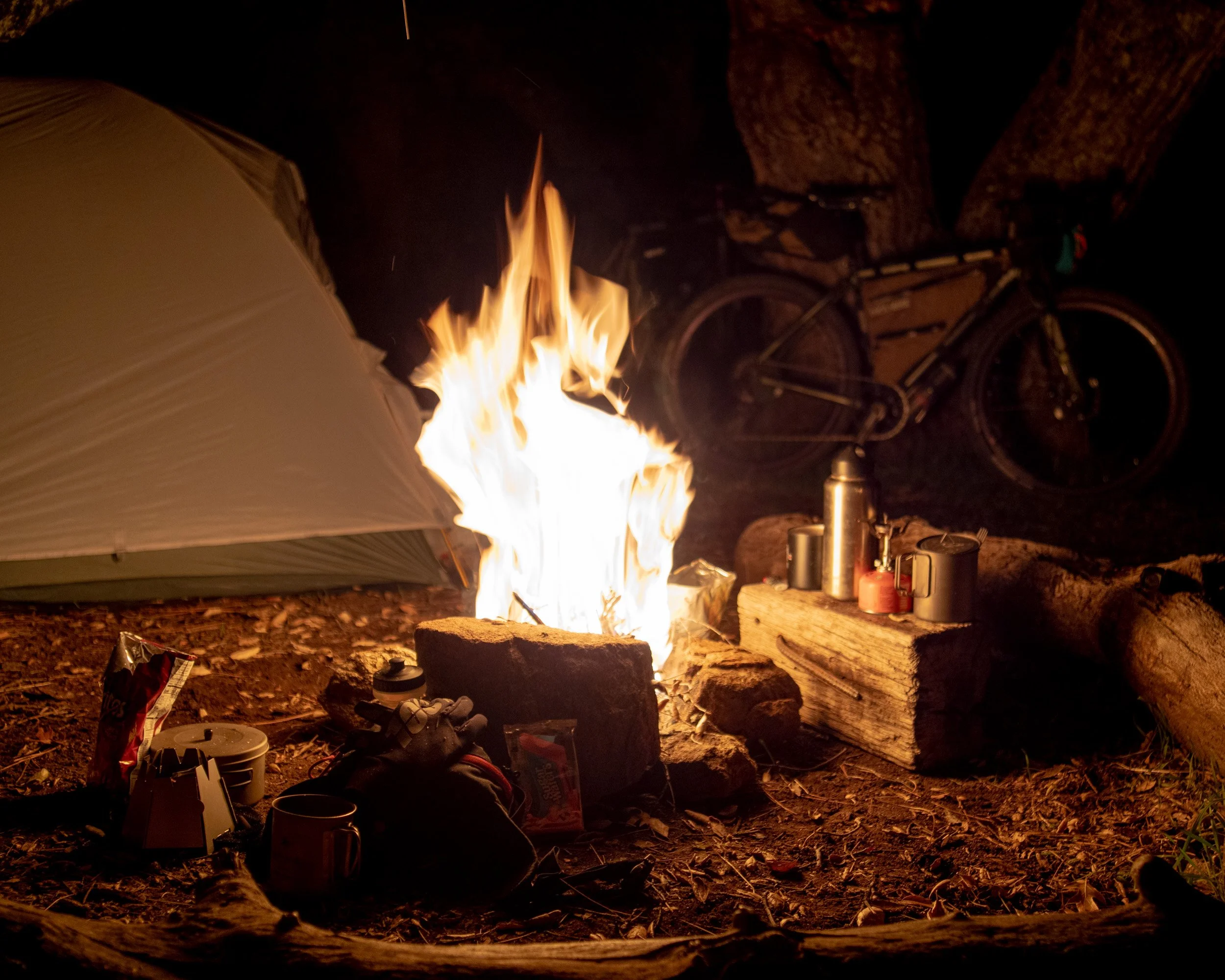 Nighttime camping scene with a campfire, a tent, a bicycle, and camping supplies on the forest ground.