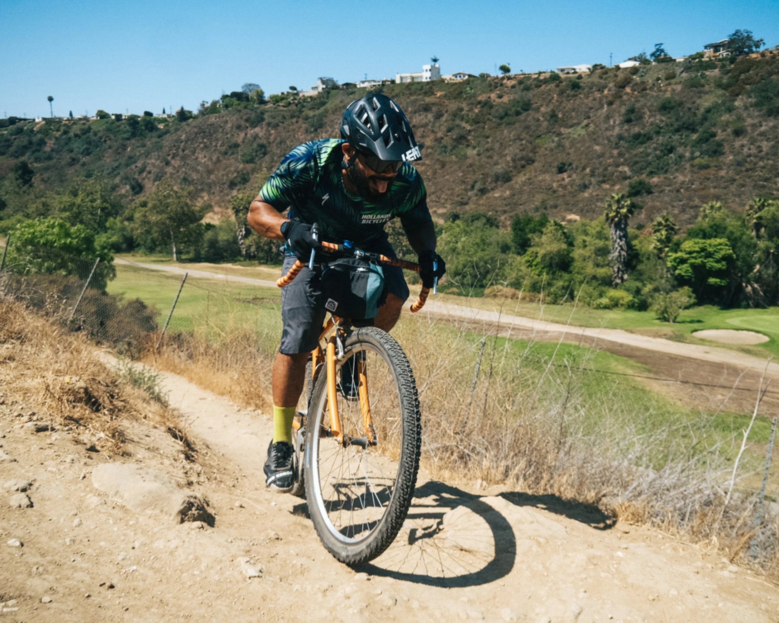 A man riding a mountain bike on a dirt trail on a sunny day, surrounded by green trees and hills.