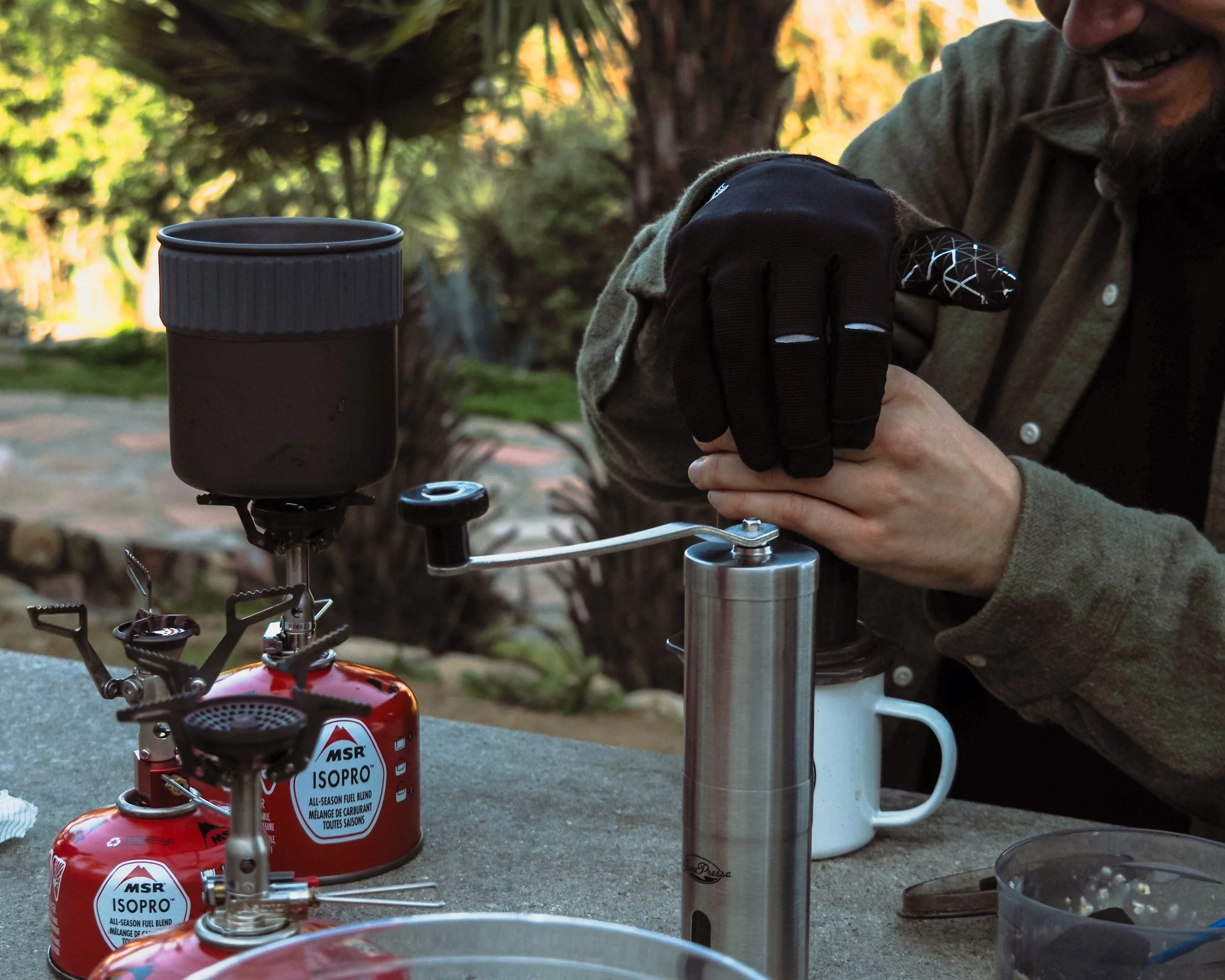 A man wearing gloves is using a hand-crank coffee grinder outdoors on a picnic table, with a coffee mug nearby. There are two small red portable gas stoves with MSR ISOPRO fuel canisters on the table, and trees with orange and green leaves in the background.