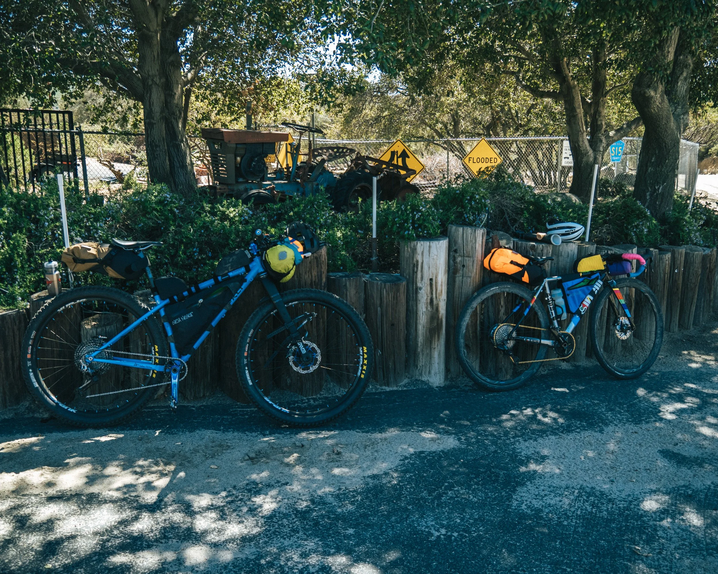 Two mountain bikes leaned against a wooden border along a paved trail. The bikes are equipped with colorful gear bags. Behind the bikes, there is green shrubbery, trees, and a fence with various signs, including one that says "Flooded" and another indicating a route. An old tractor is also visible among the trees and shrubs.