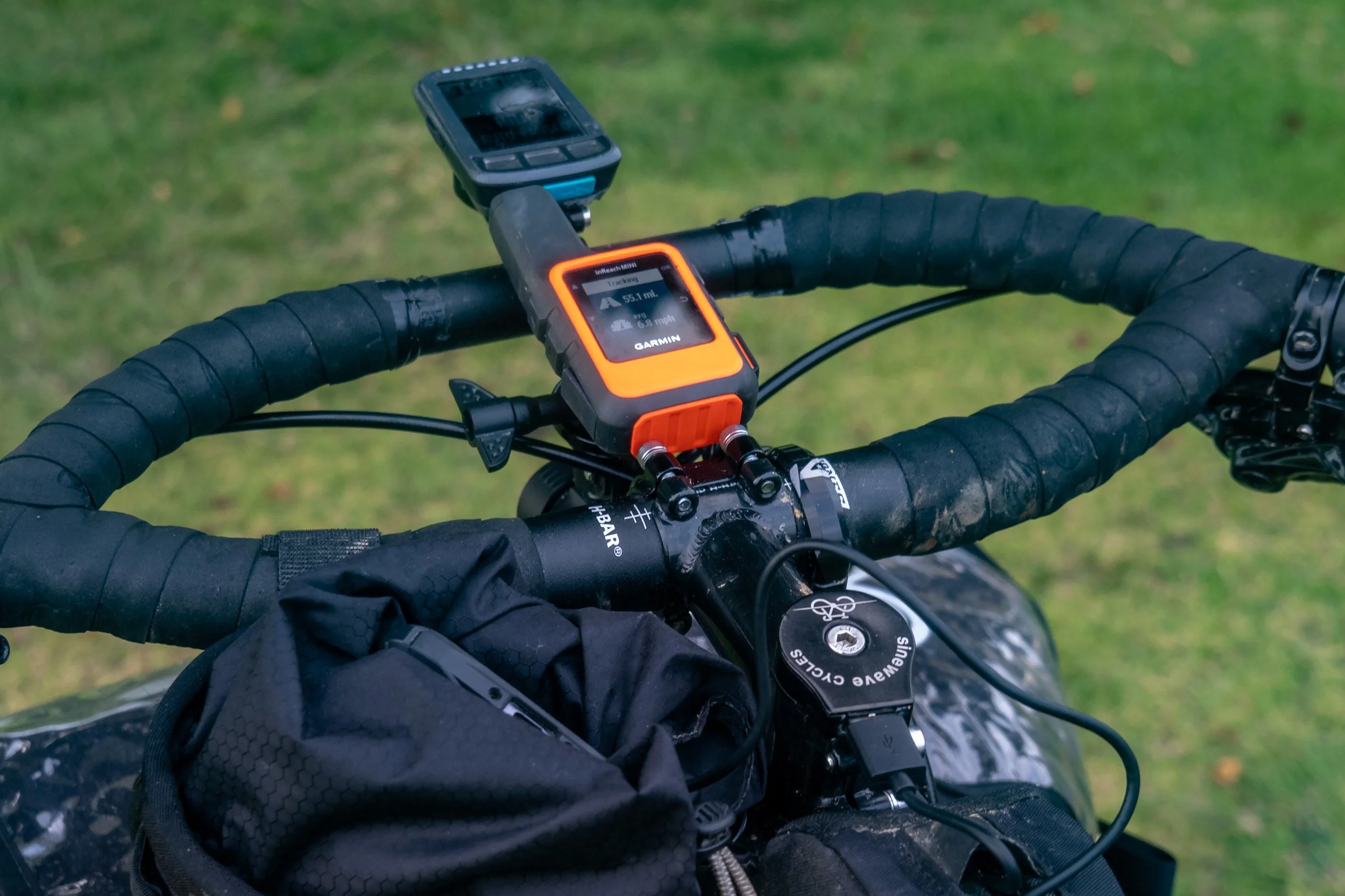 Close-up of a bicycle handlebar with electronic cycling devices attached, including a Garmin GPS unit and a cycling computer, against a blurred grassy background.