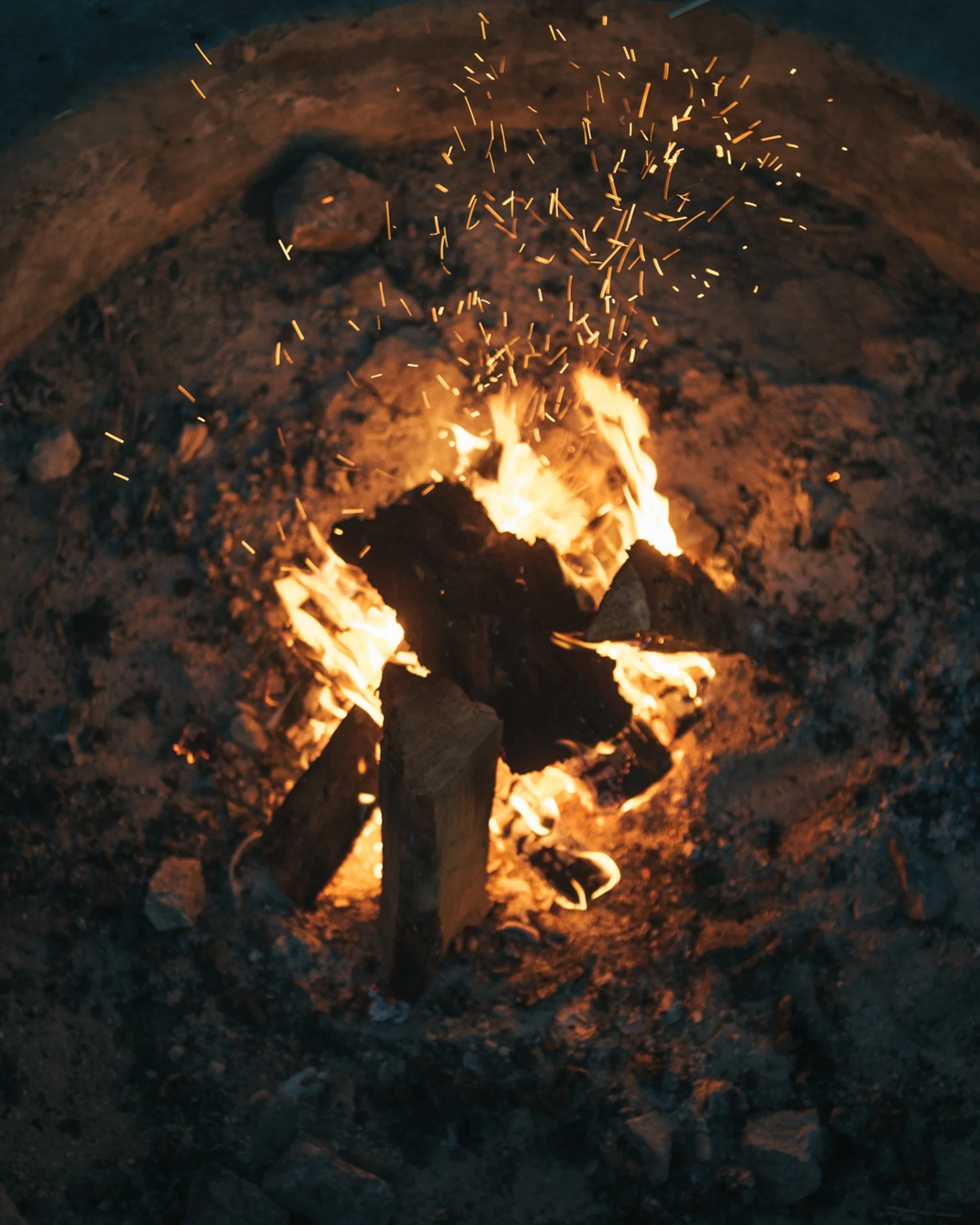 A small campfire burning on dirt ground with sparks flying upward.
