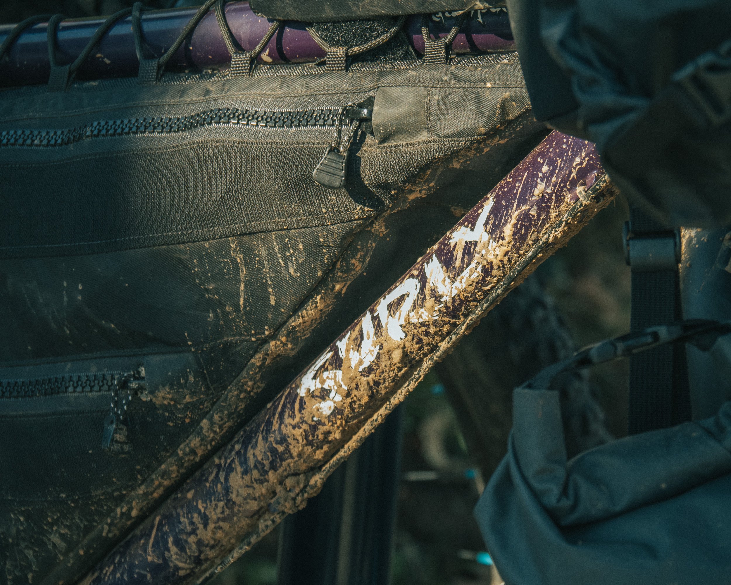 Close-up of a muddy bicycle with the brand name 'SURGE' partially visible on the frame, next to a black backpack with zippers.