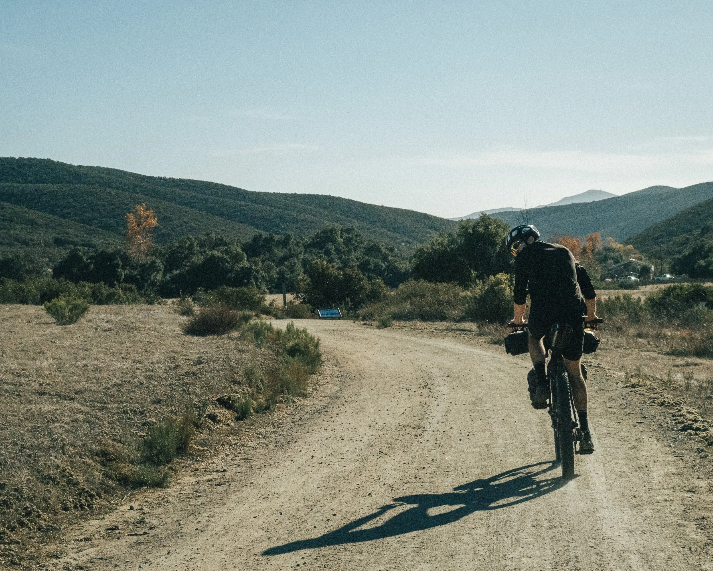 A person riding a mountain bike on a dirt trail through a hilly landscape with trees and shrubs, under a clear sky.