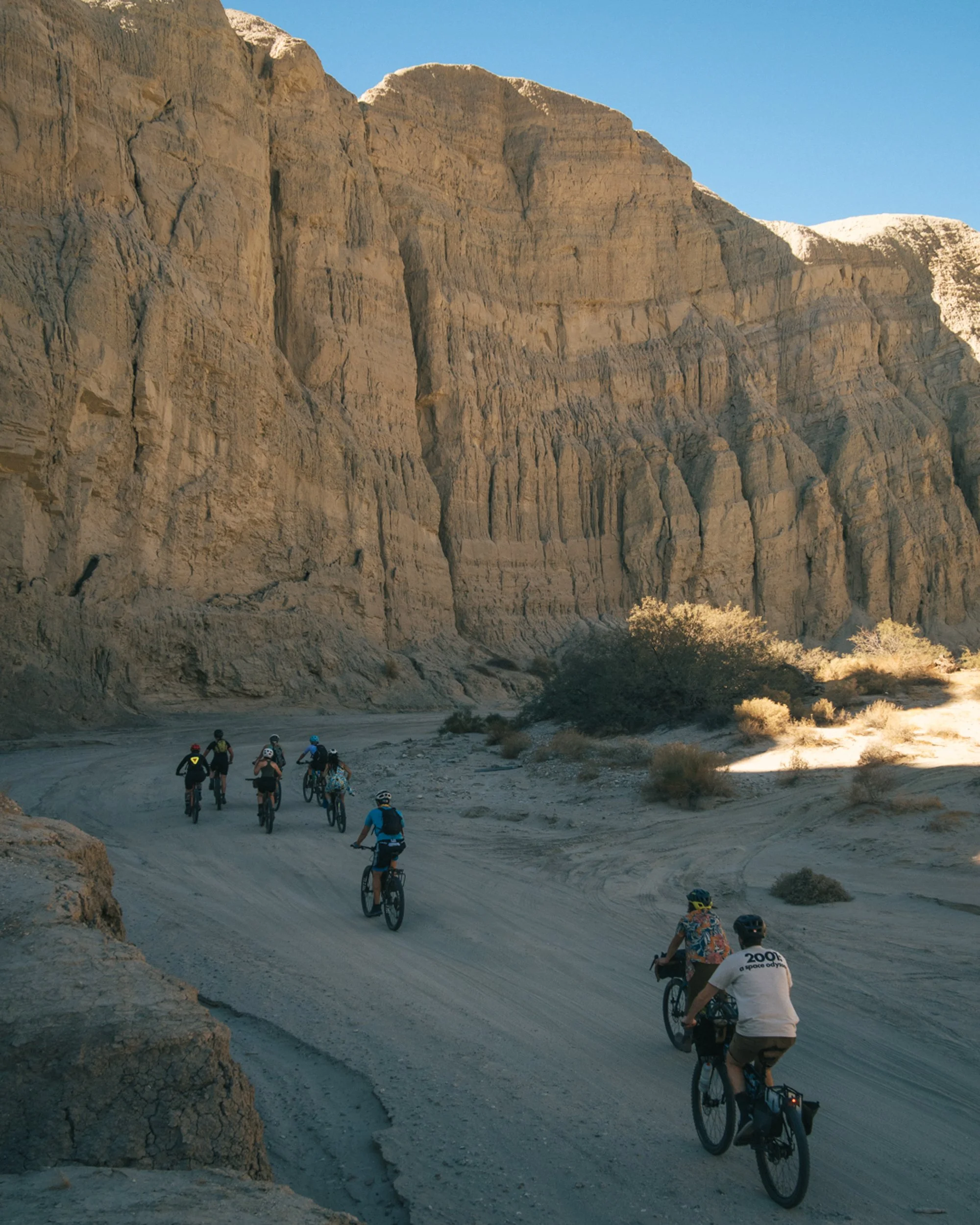 Group of cyclists riding on a dirt trail through a desert canyon with tall rocky cliffs and sparse vegetation.