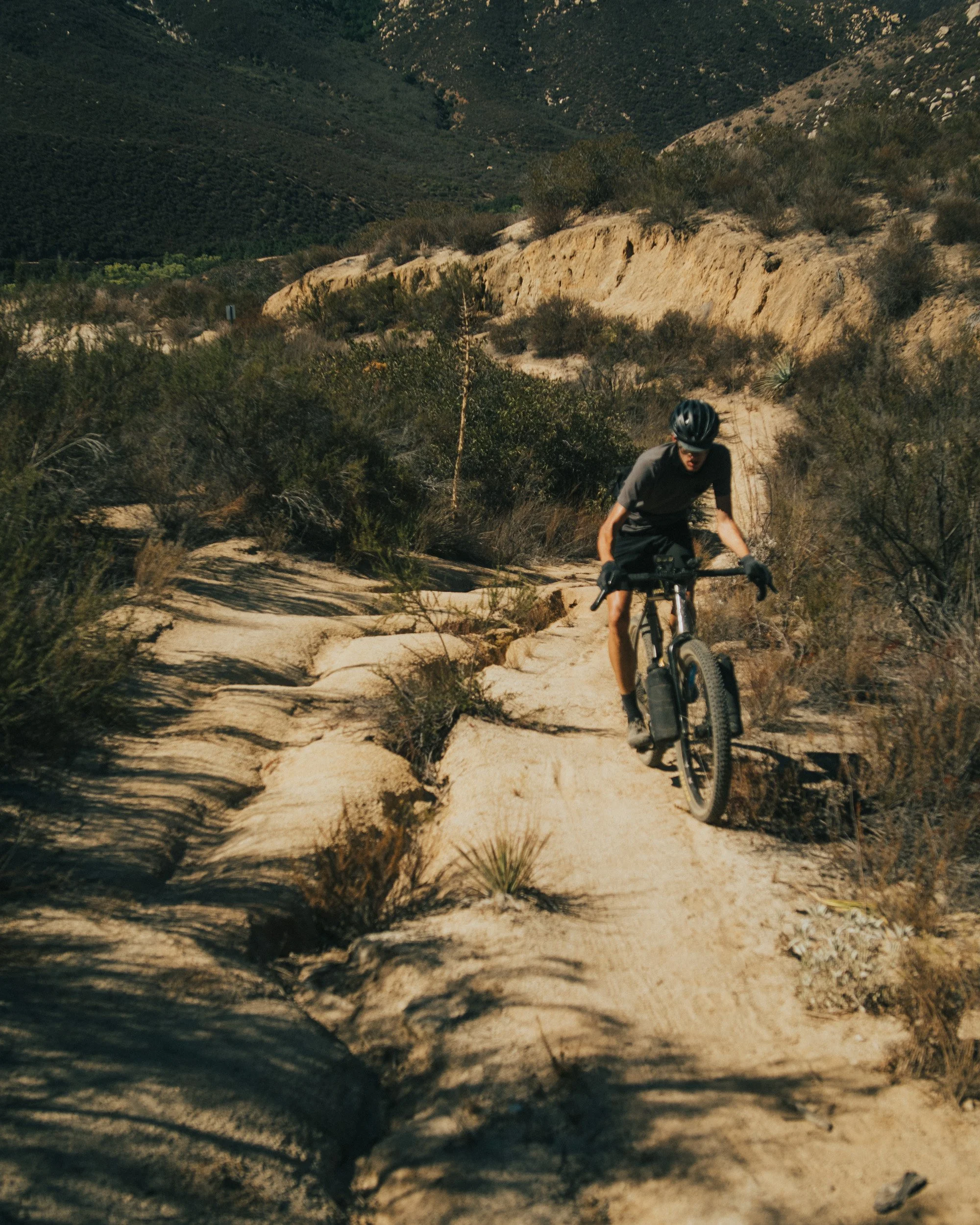 A person riding a mountain bike on a rocky trail in a desert landscape with shrubs and hills in the background.