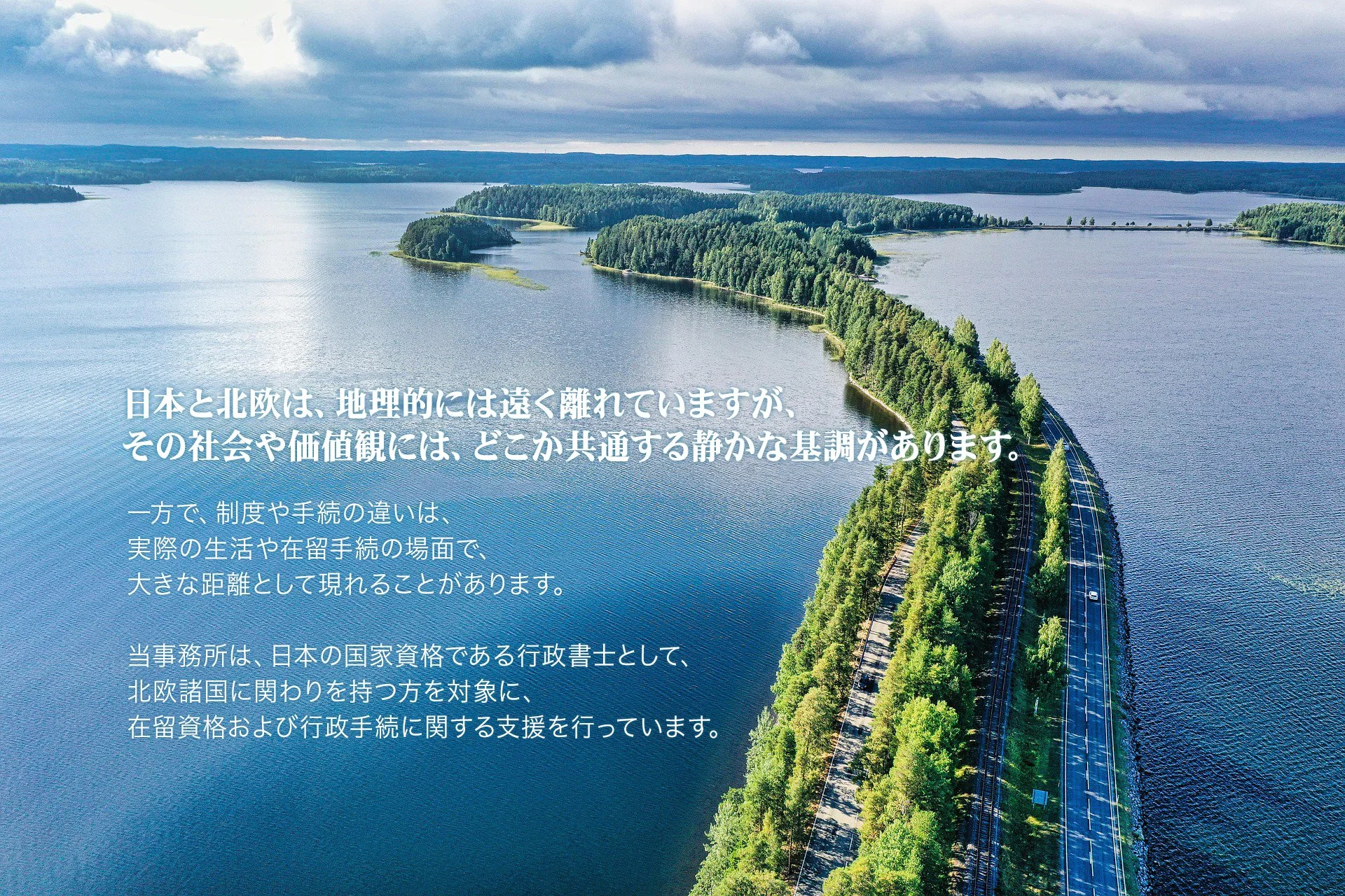 Aerial view of a large body of water with an island covered in dense green trees, and a railway track running along the edge of the island under a cloudy sky.