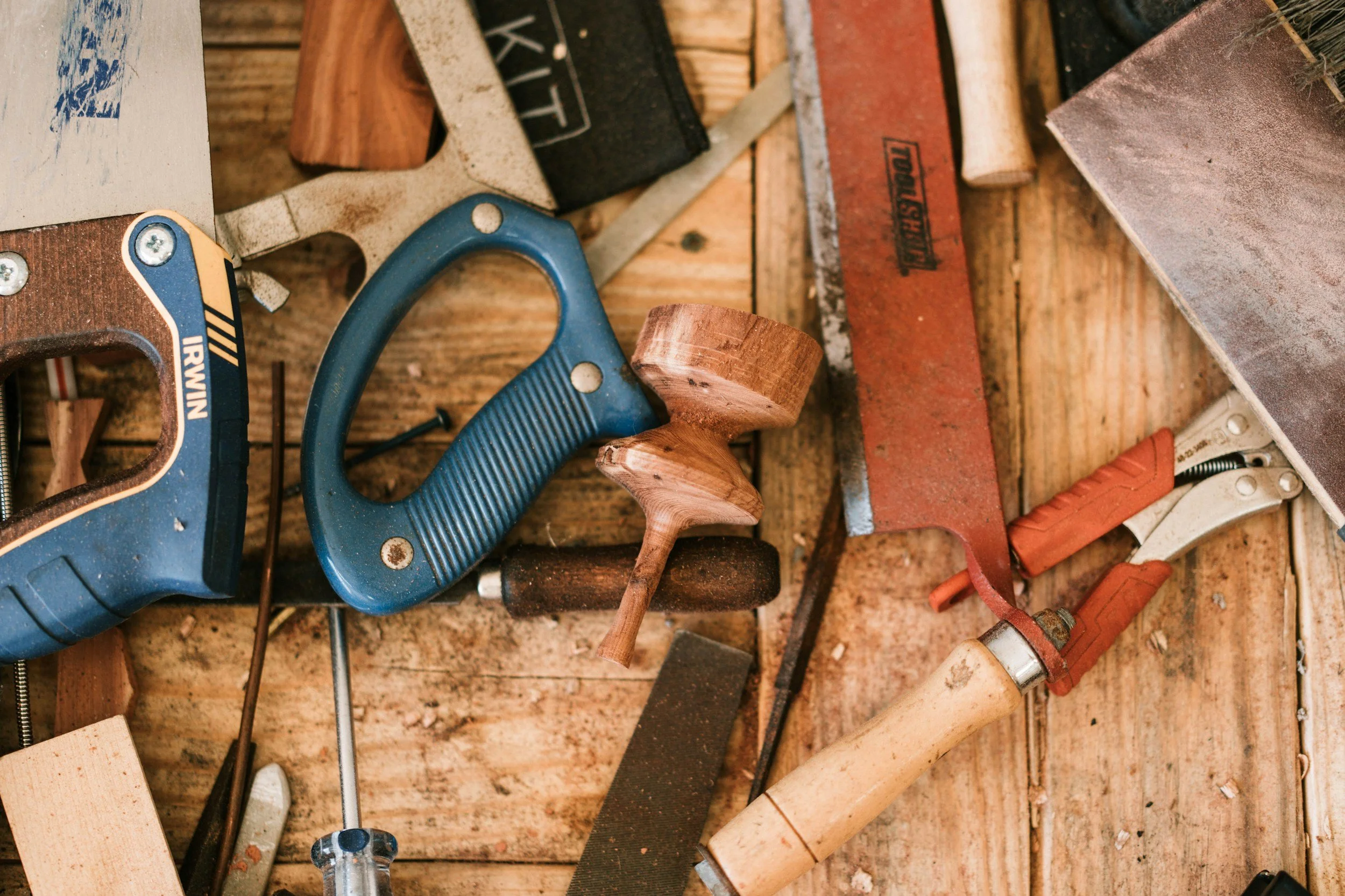A scattered assortment of woodworking tools on a wooden surface, including a blue hacksaw, a wooden mallet, a red metal file, a clamp, rasps, screwdrivers, and other small hand tools.