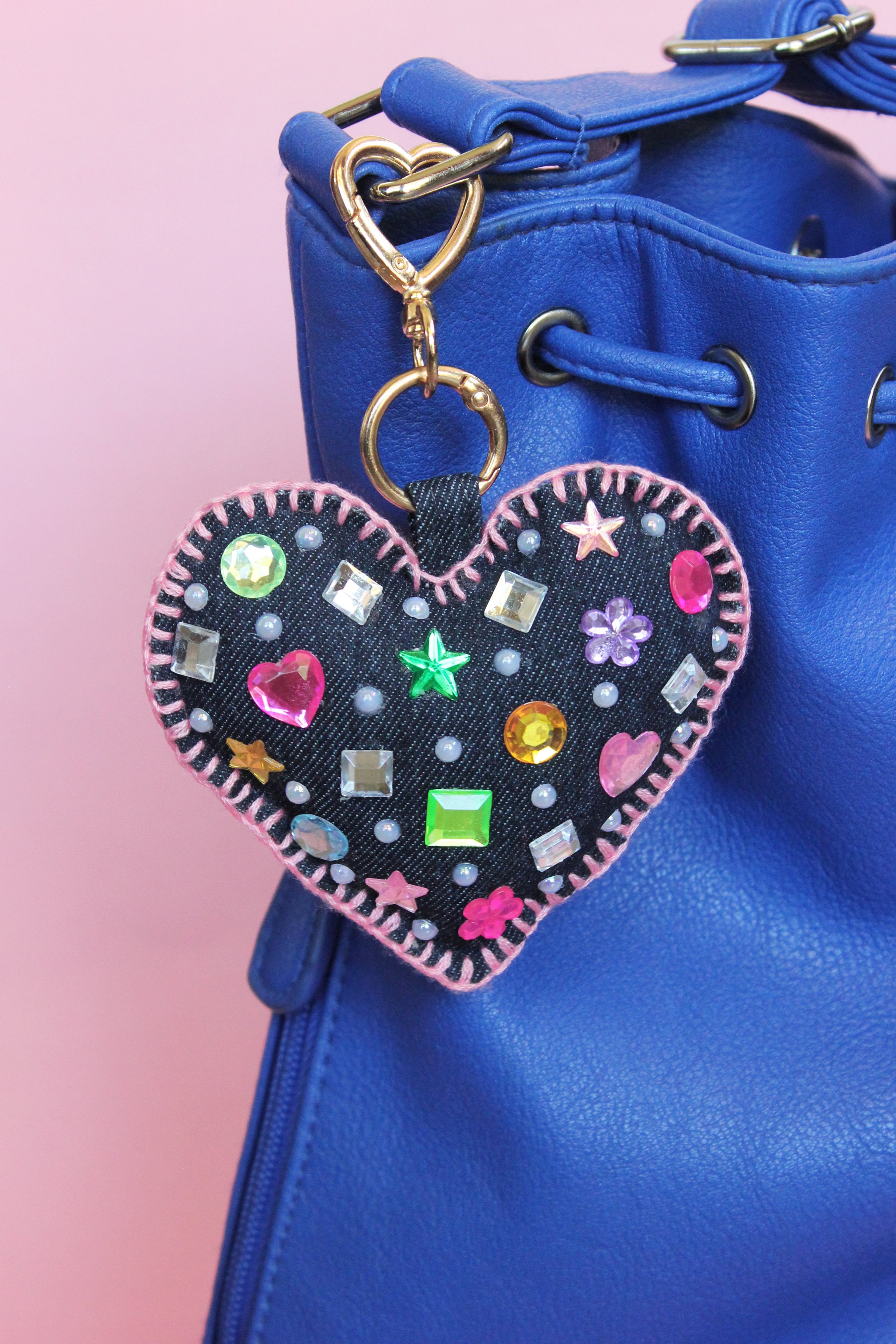 A blue handbag with gold hardware and a decorated heart-shaped keychain with colorful rhinestones and beads attached to the handle.