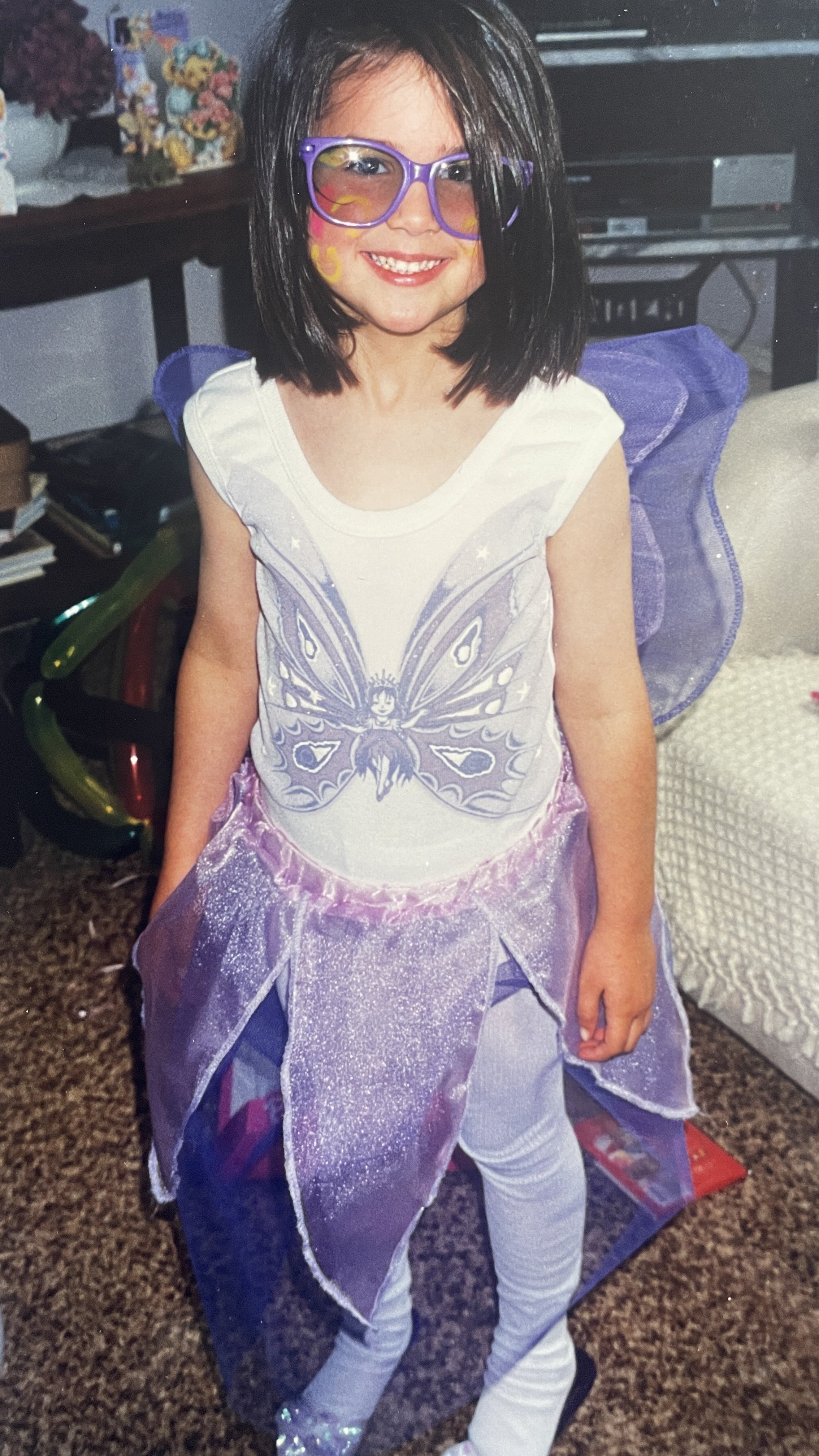 A young girl dressed as a fairy with purple wings, a butterfly graphic on her shirt, purple glasses, and glittery purple skirt, smiling indoors.