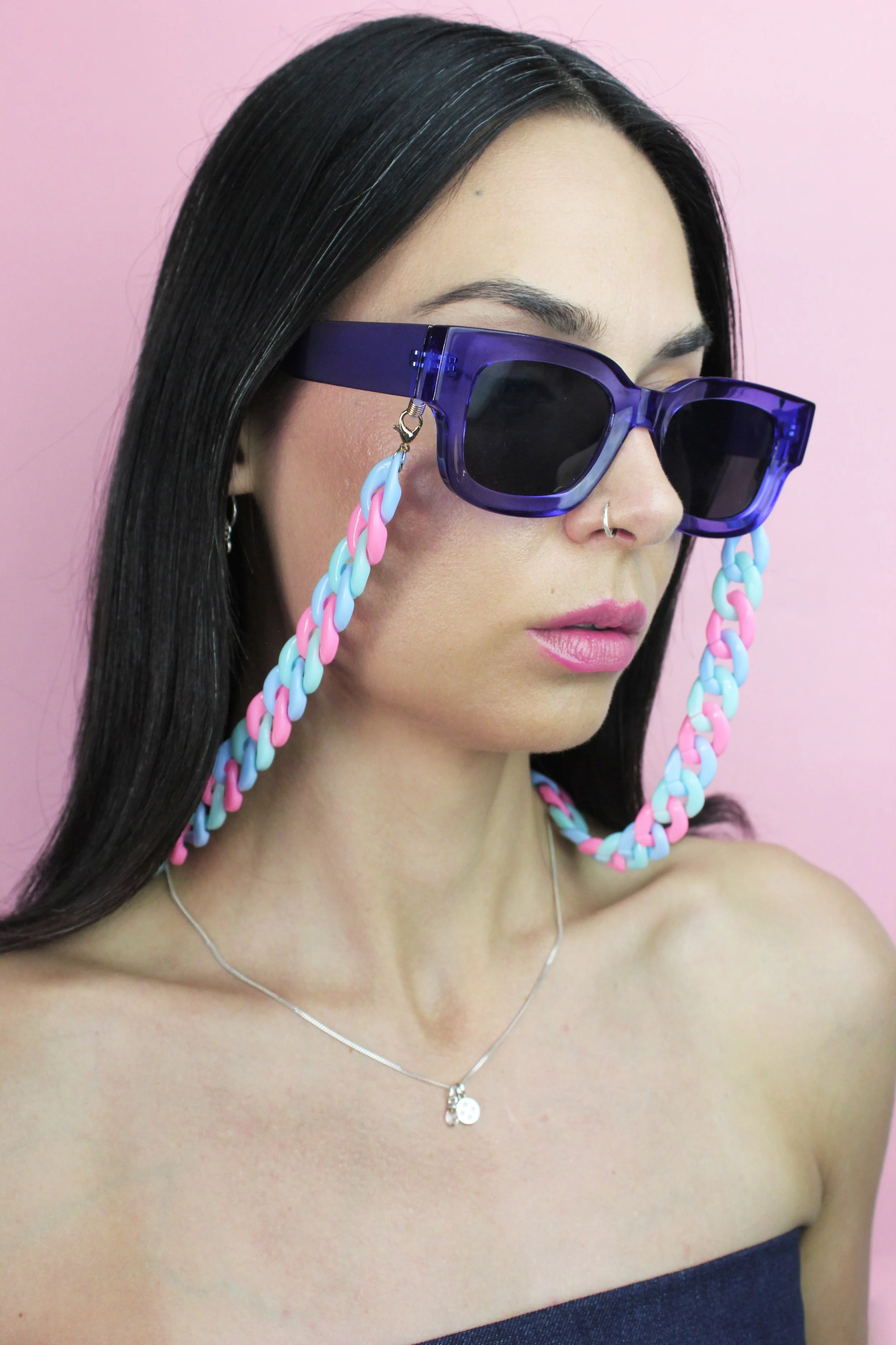 Woman with long dark hair wearing large purple sunglasses with pastel-colored chain attached, pink lipstick, silver necklace, and a strapless top against a pink background.