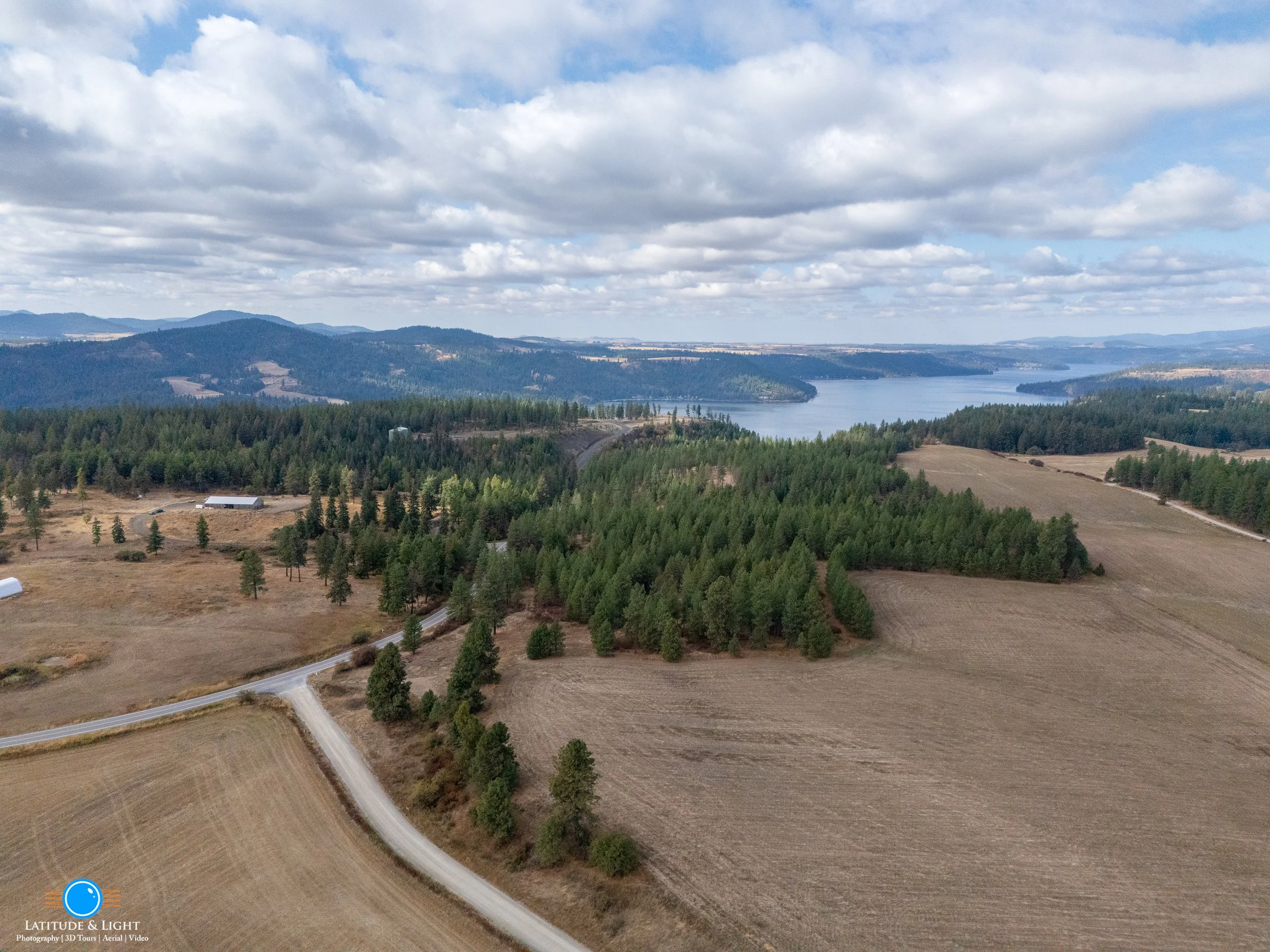 Aerial view of a rural landscape in North Idaho with fields, forests, a winding road, a river or lake, and distant mountains under a partly cloudy sky.