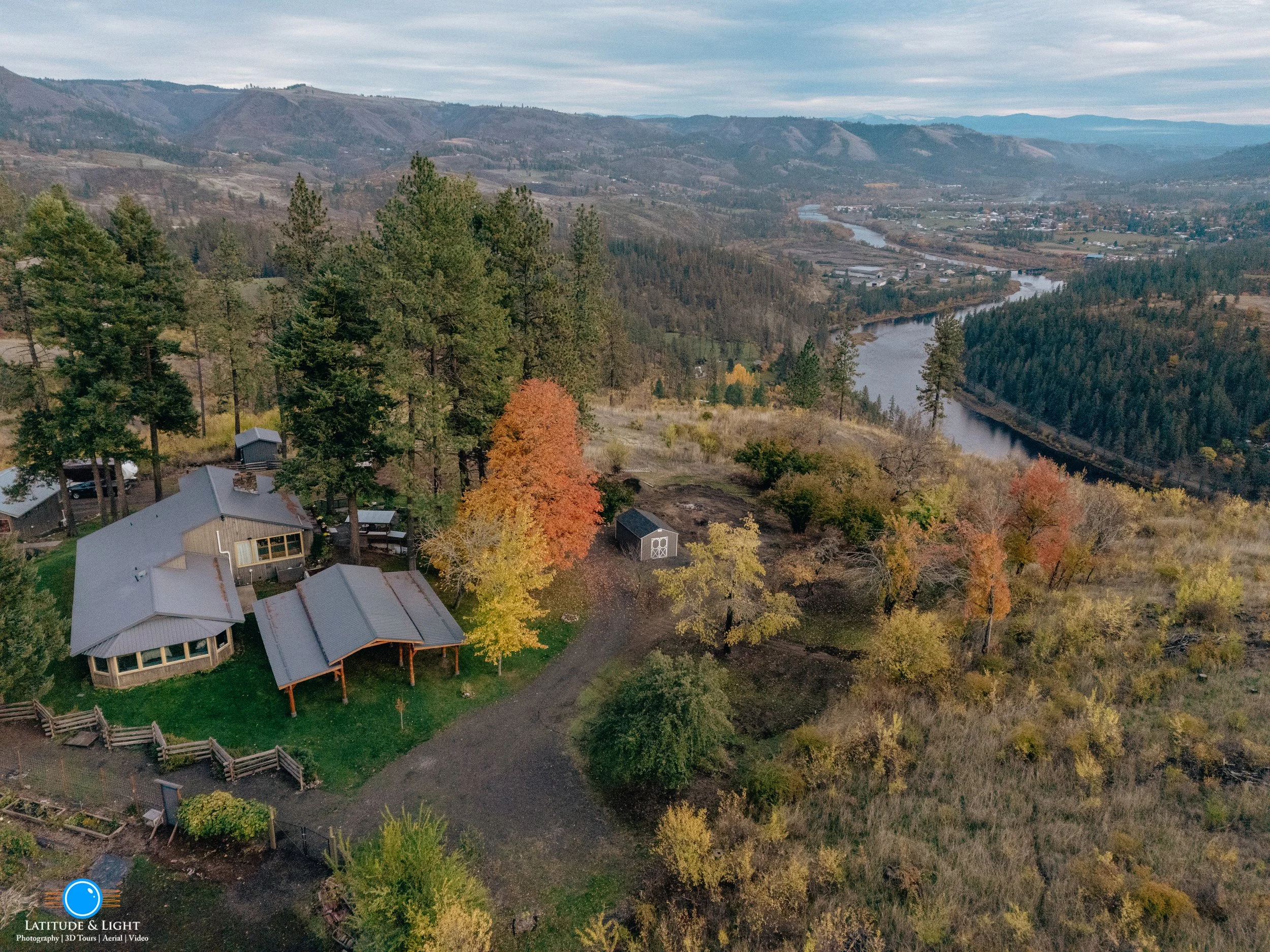 Aerial view of a North Idaho house urrounded by tall pine trees and autumn-colored trees, near a river winding through a valley with mountains in the distance.