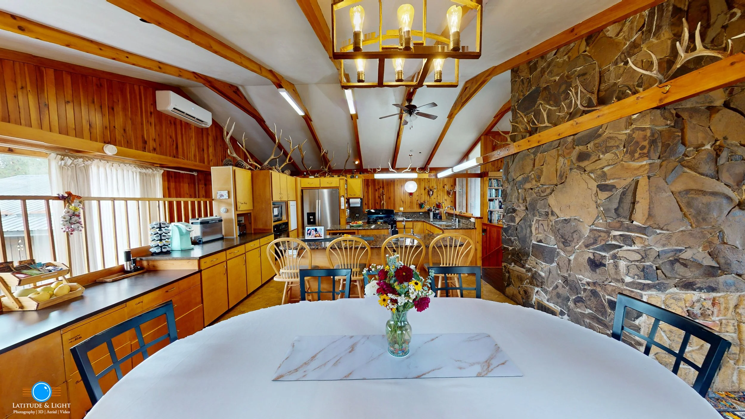 Interior of a rustic kitchen and dining area in a rural Montana wedding ranch with wooden walls and ceiling beams, featuring a large stone wall with mounted antlers, a round table with a white tablecloth and a flower arrangement.