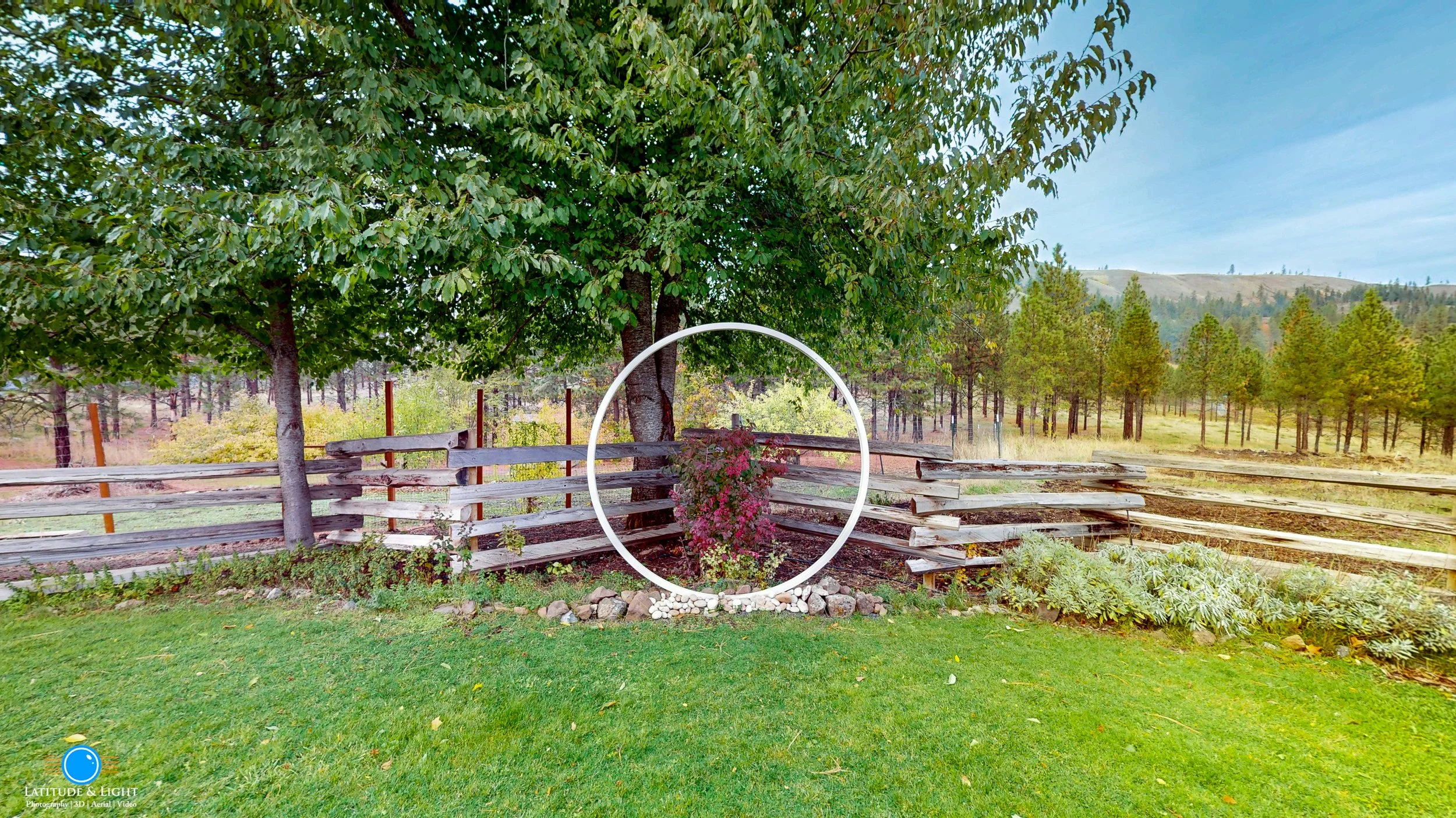 A ceremony area at a Montana wedding ranch sits in grassy yard with a large leafy tree, a wooden fence, and a group of pine trees in the background. There is a decorative white circle in the center around a small bush with pink flowers.
