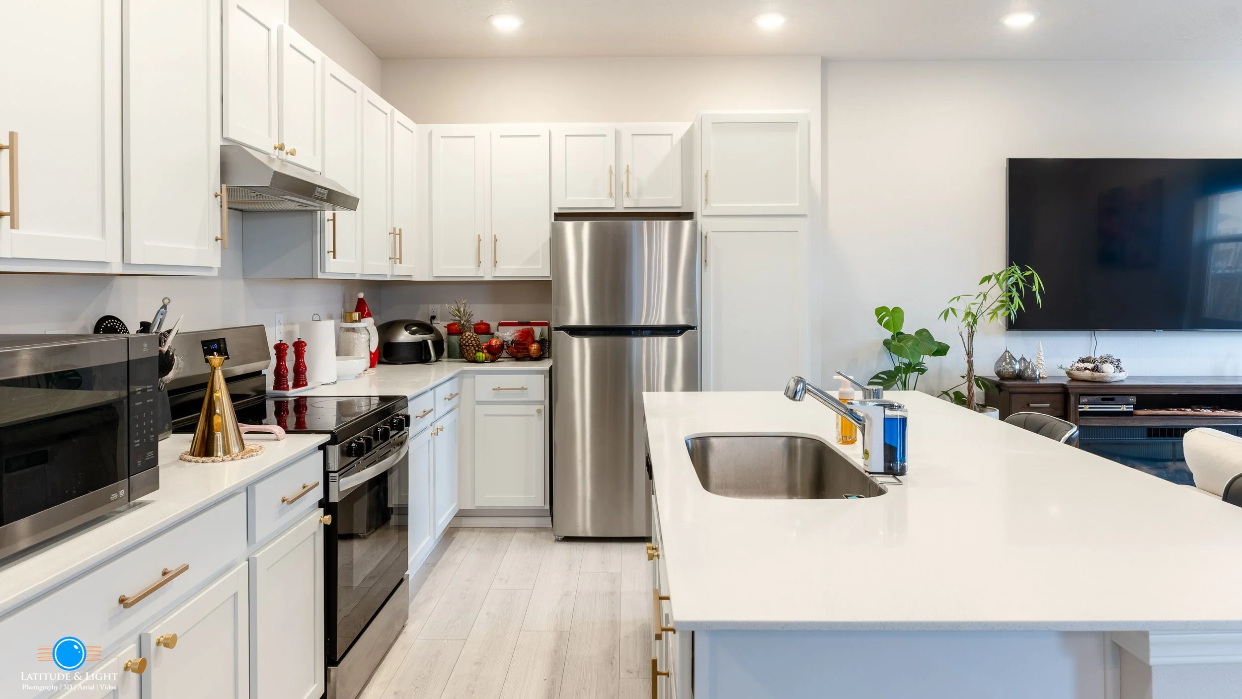 Modern kitchen with white cabinets, stainless steel refrigerator, black stove, microwave, and a kitchen island with a sink. Decor includes fruit, plants, and holiday-themed decorations.