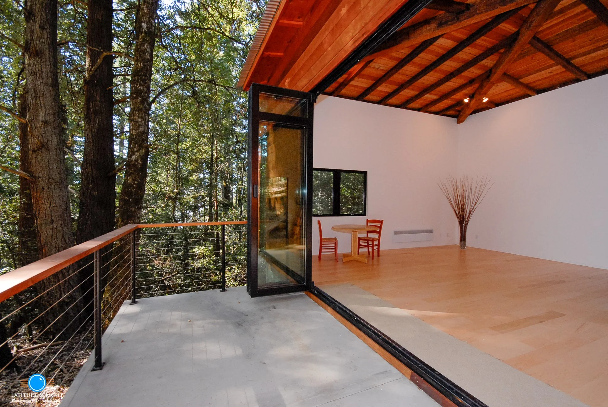 Interior of a Los Gatos home with a large open sliding glass door leading to a balcony with a forest view, wood ceiling, minimal furniture, and a wooden floor.