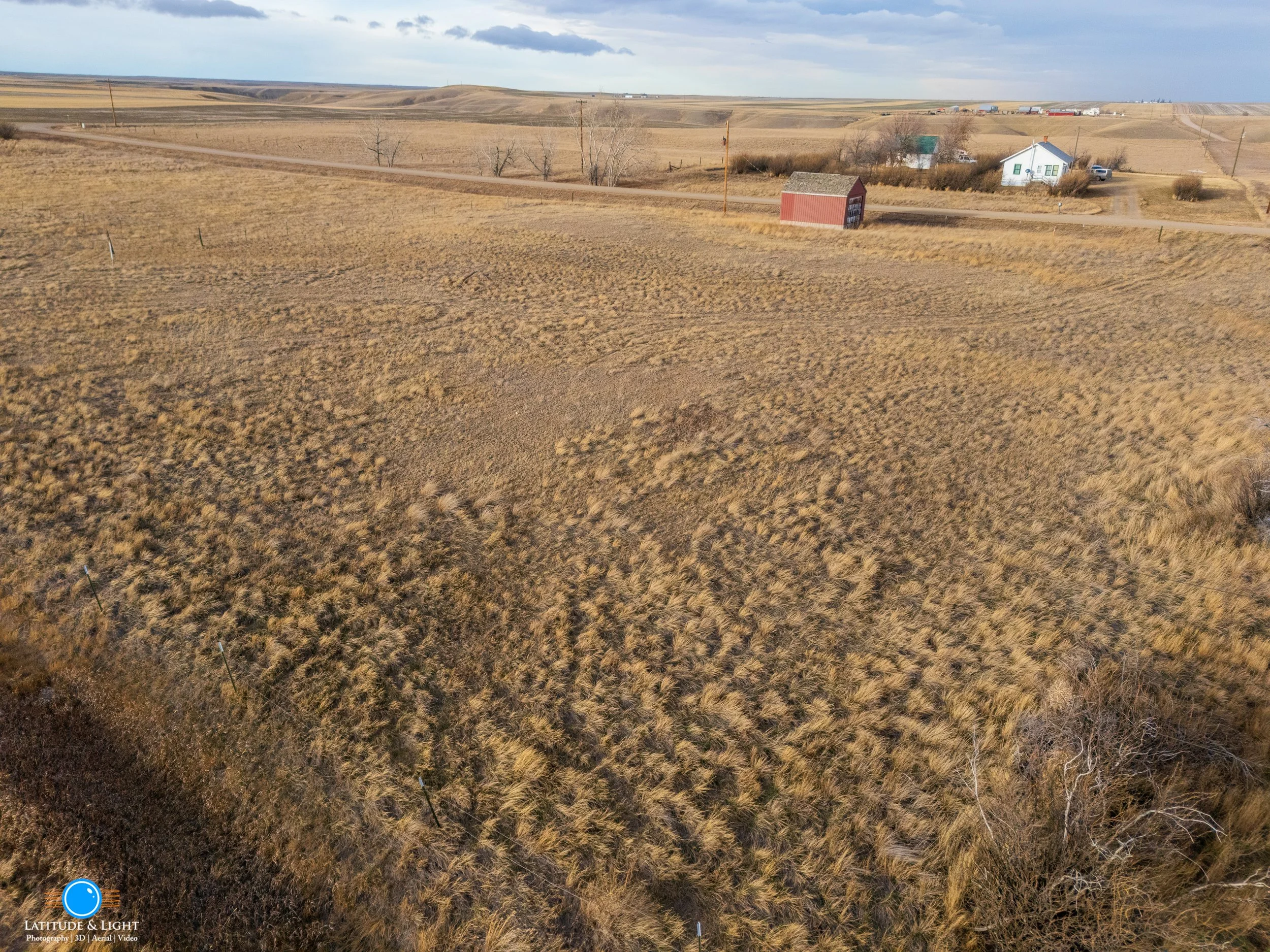 North Idaho land: Wide view of rural farmland with dry grass, a small dirt road, scattered trees, and farm buildings in the distance under a partly cloudy sky.
