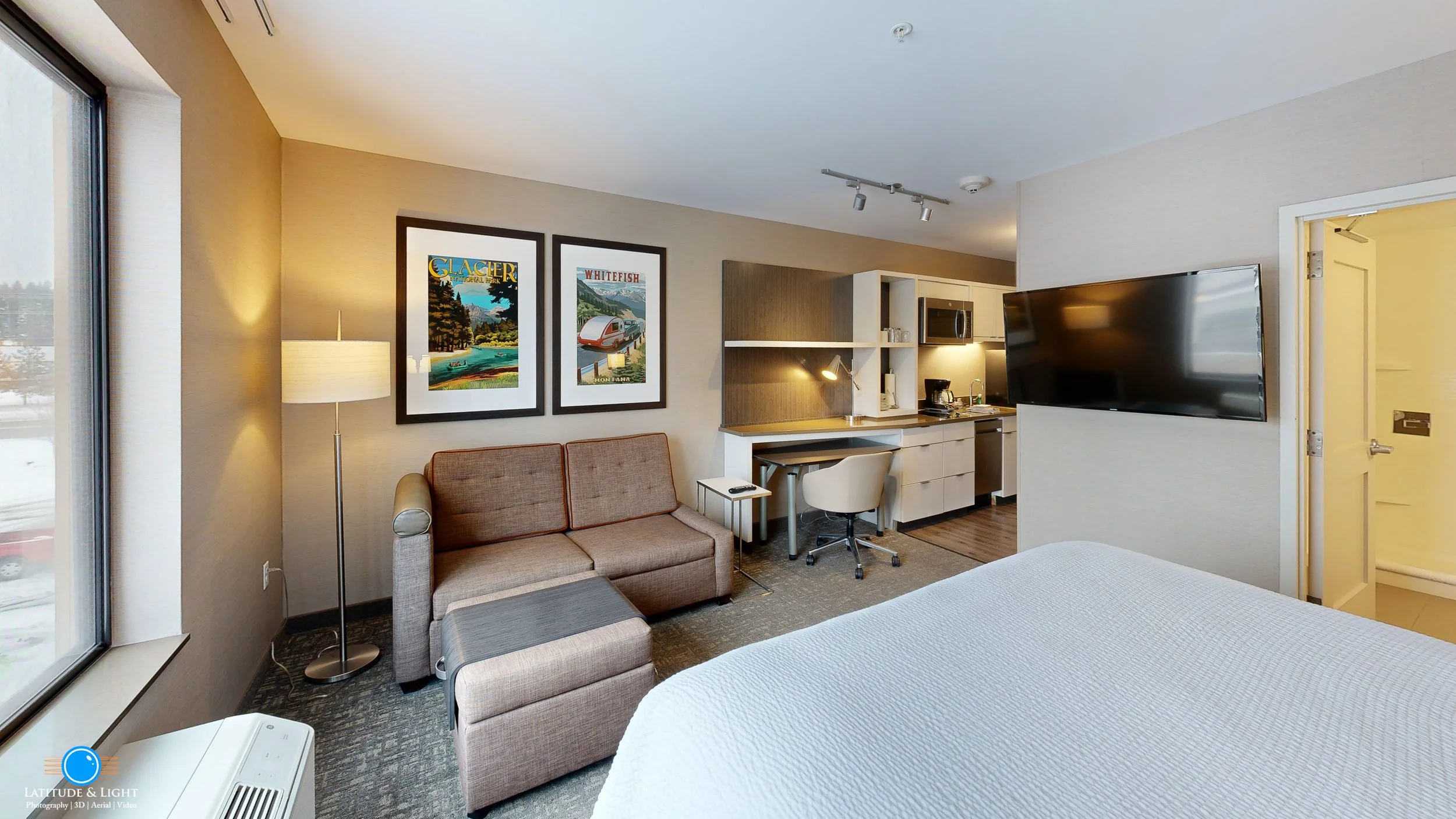 Hotel room with bed, sofa, wall-mounted TV, small kitchen area, window showing snowy outdoors, artwork on wall, and desk with chair.