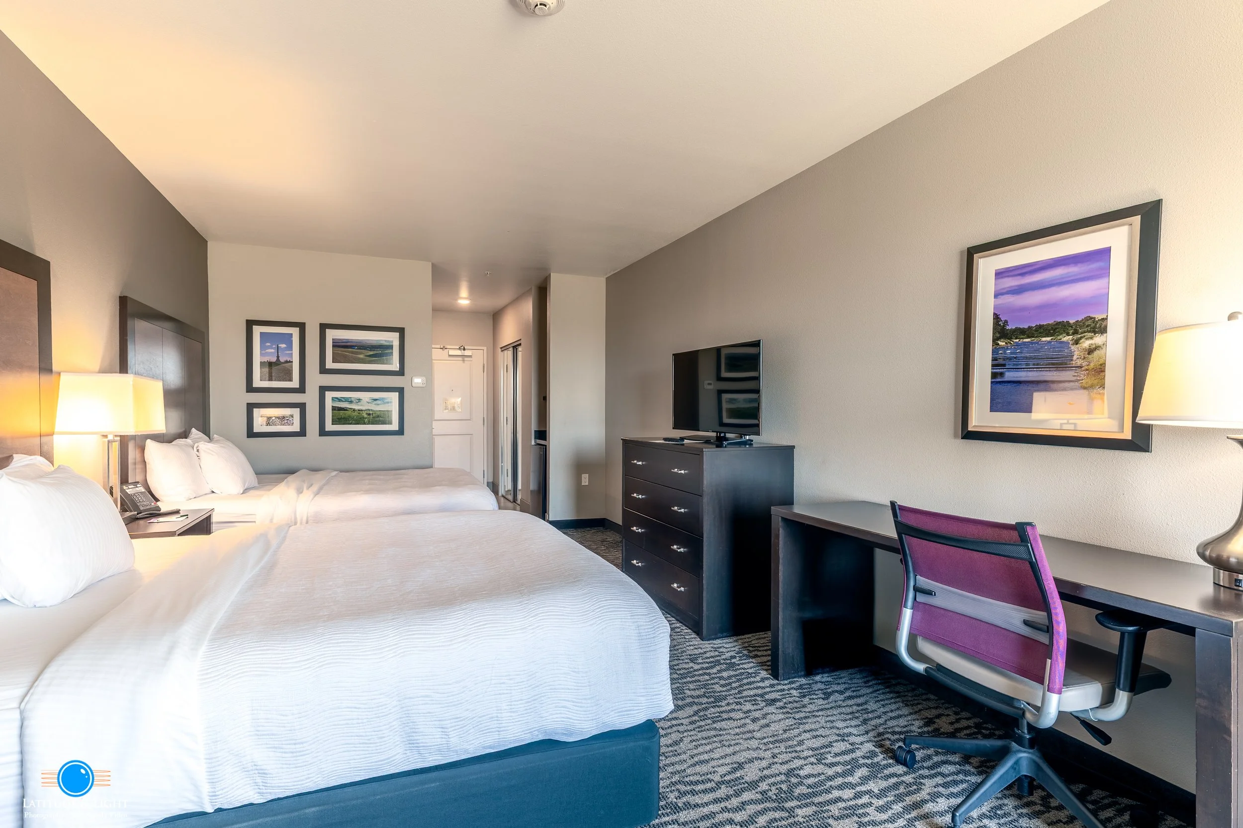 A hotel room with two queen beds, a television on a black dresser, a desk with a purple chair, wall art, and a lamp, with a carpeted floor and an entry door in the background.
