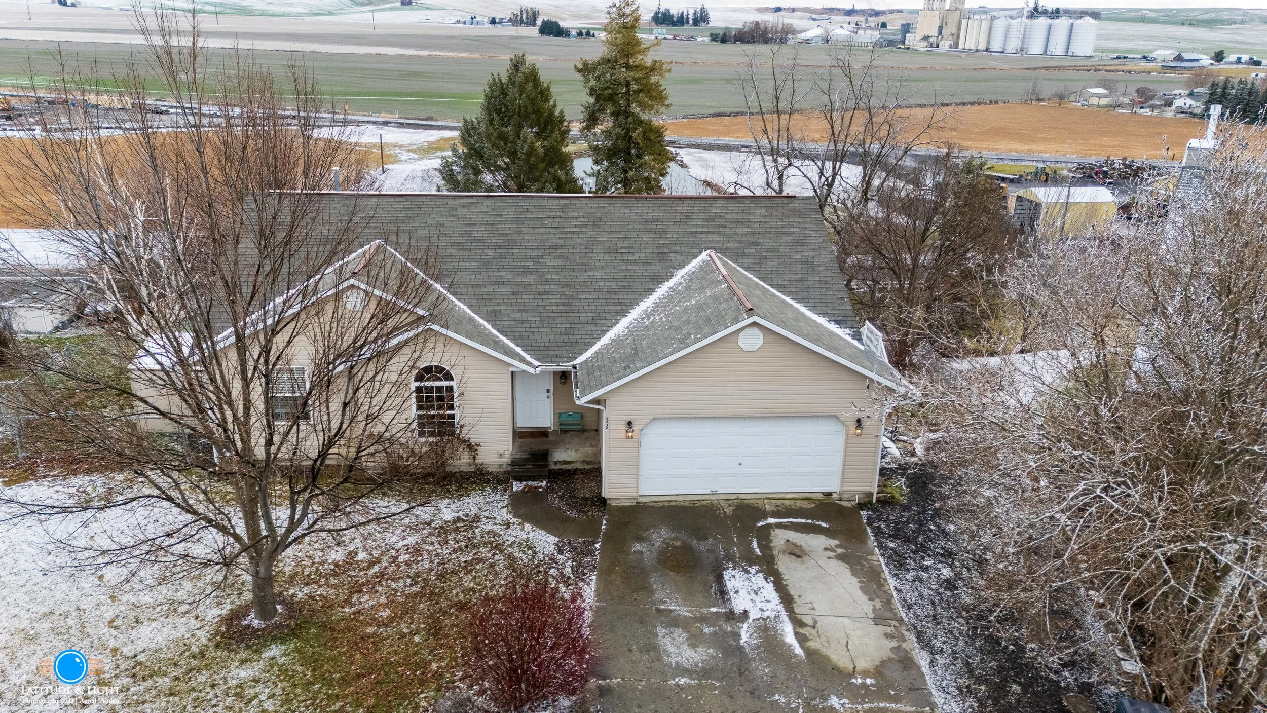 Aerial view of a single-story house in Moscow, Idaho with a gray roof and beige siding, surrounded by leafless trees and patches of snow on the ground, with a concrete driveway leading to a white garage door and a small front porch.
