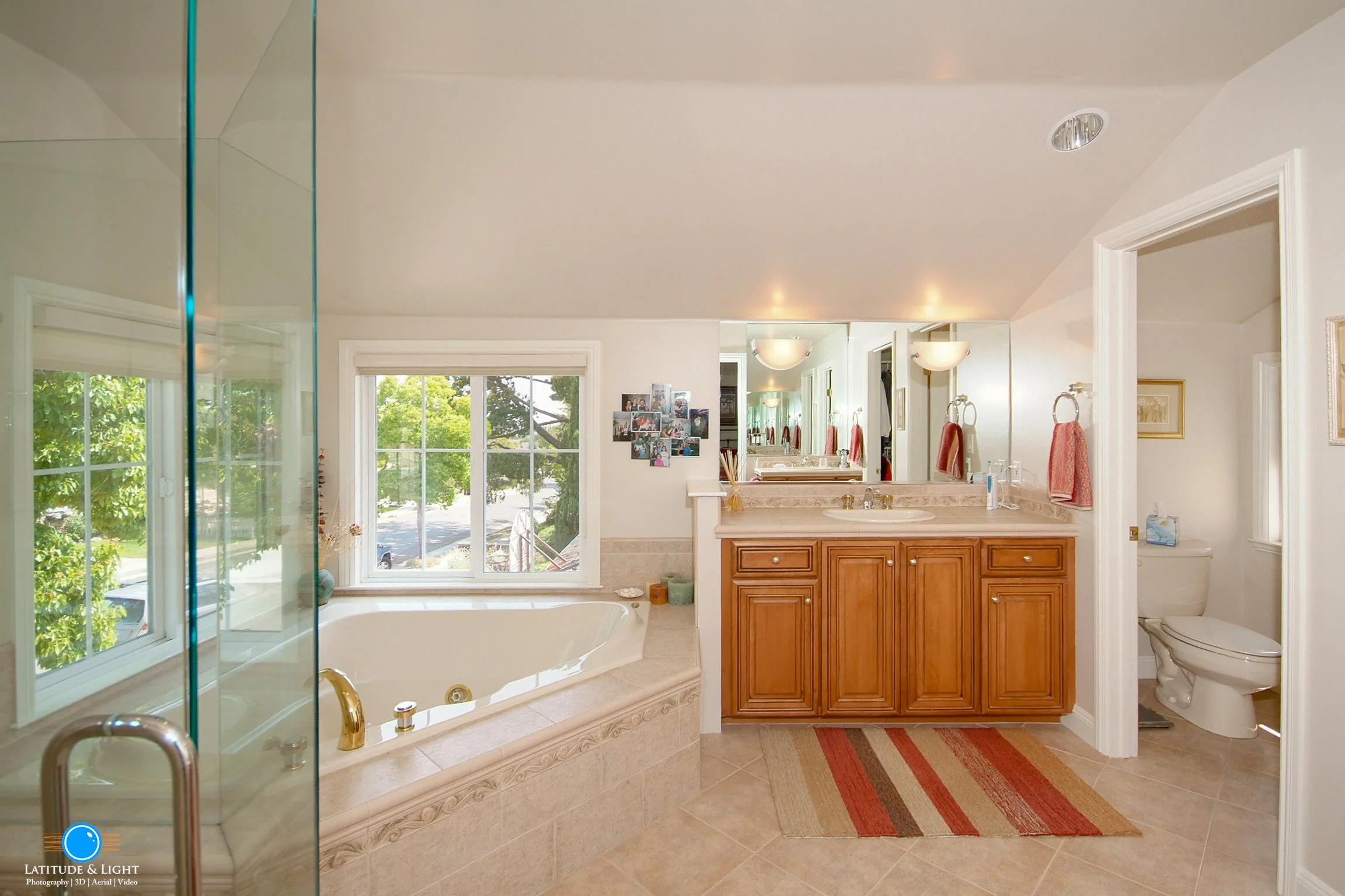Bathroom with large window over a bathtub, wooden vanity with mirror, and separate toilet room, with beige tiles and a striped rug.