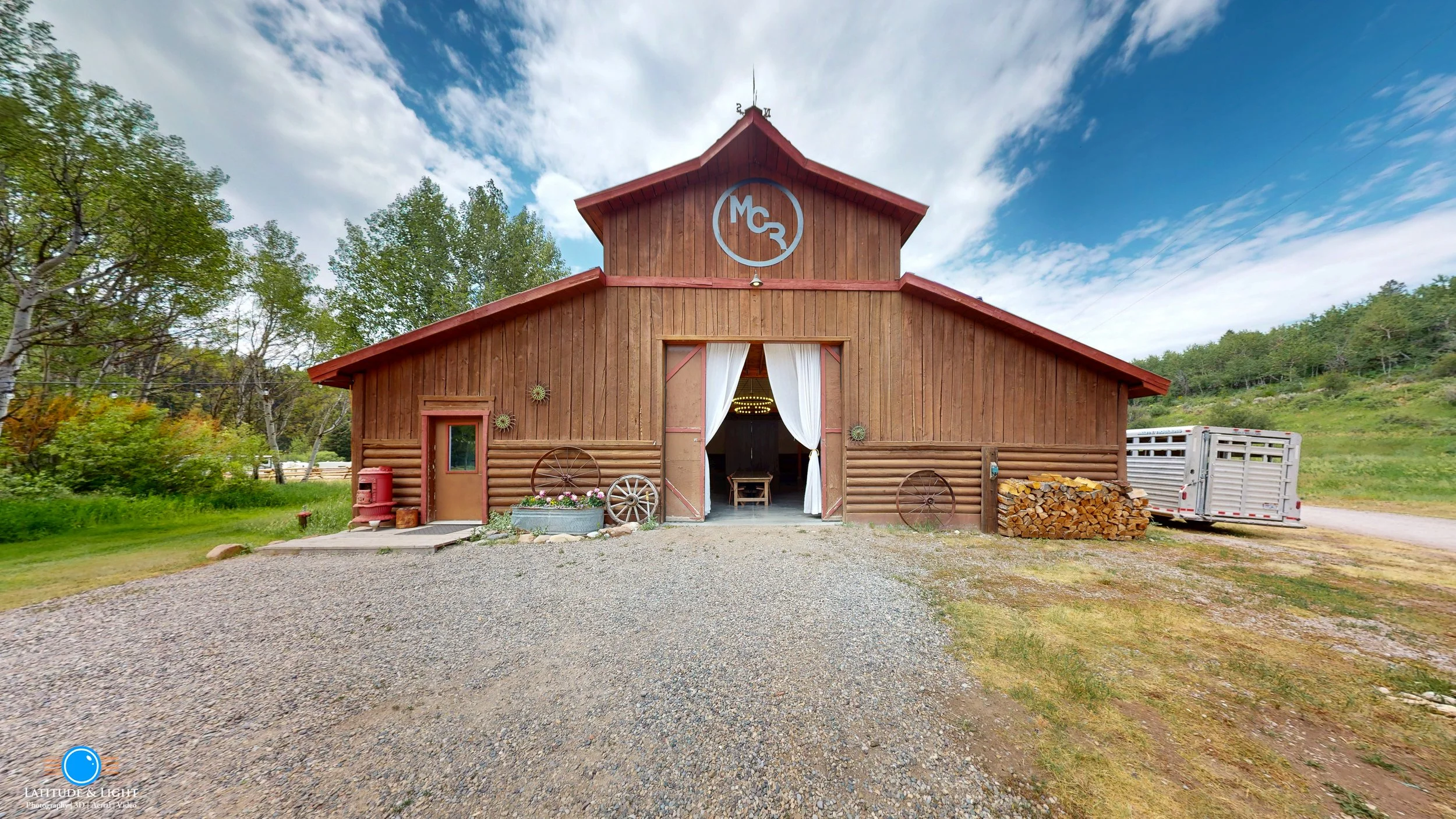A rustic wooden at a wedding ranch in Victor, Idaho with a red roof, white curtains at the entrance, surrounded by trees and greenery, with a gravel driveway in front and a trailer and firewood on the side.