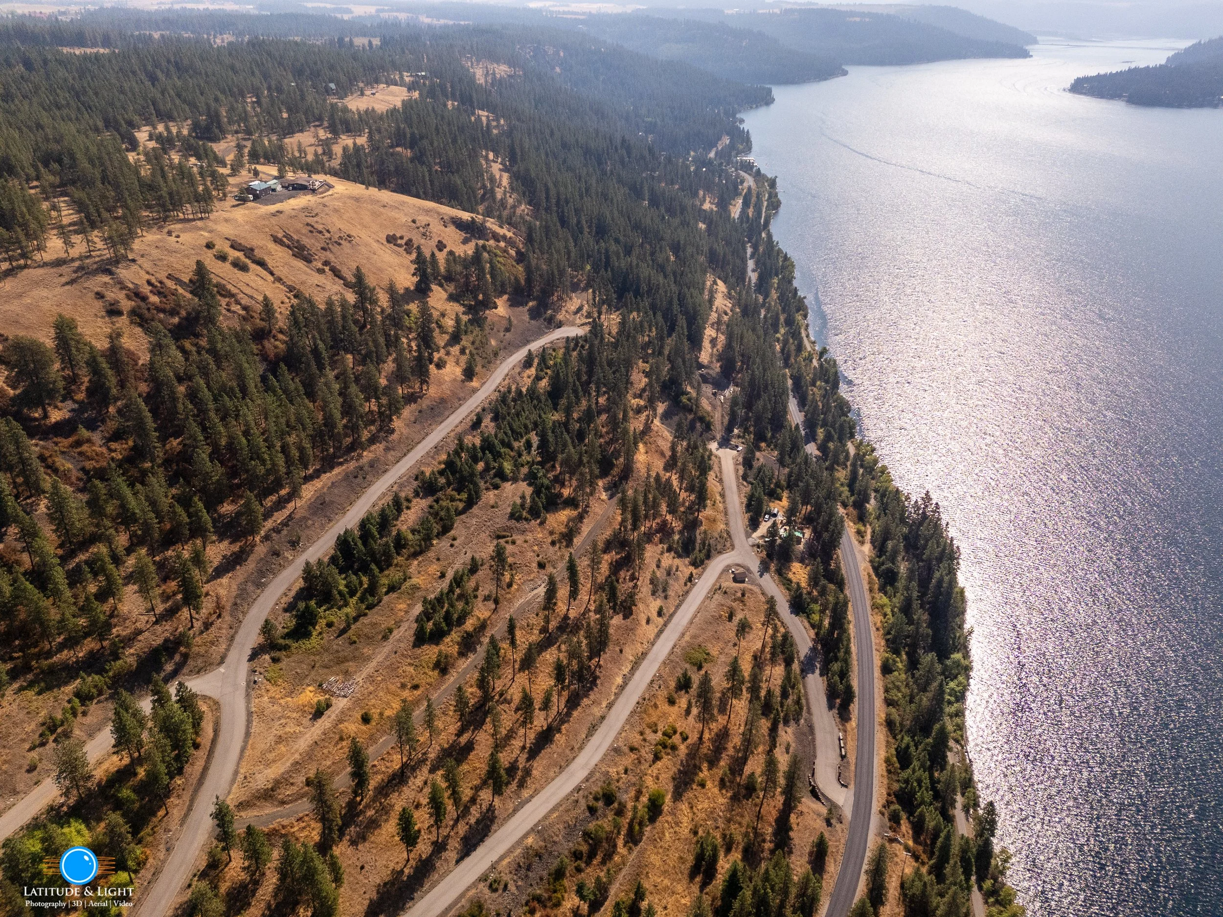 Aerial view of a winding road along a forested hillside beside a large body of water with distant mountains and cloudy sky.