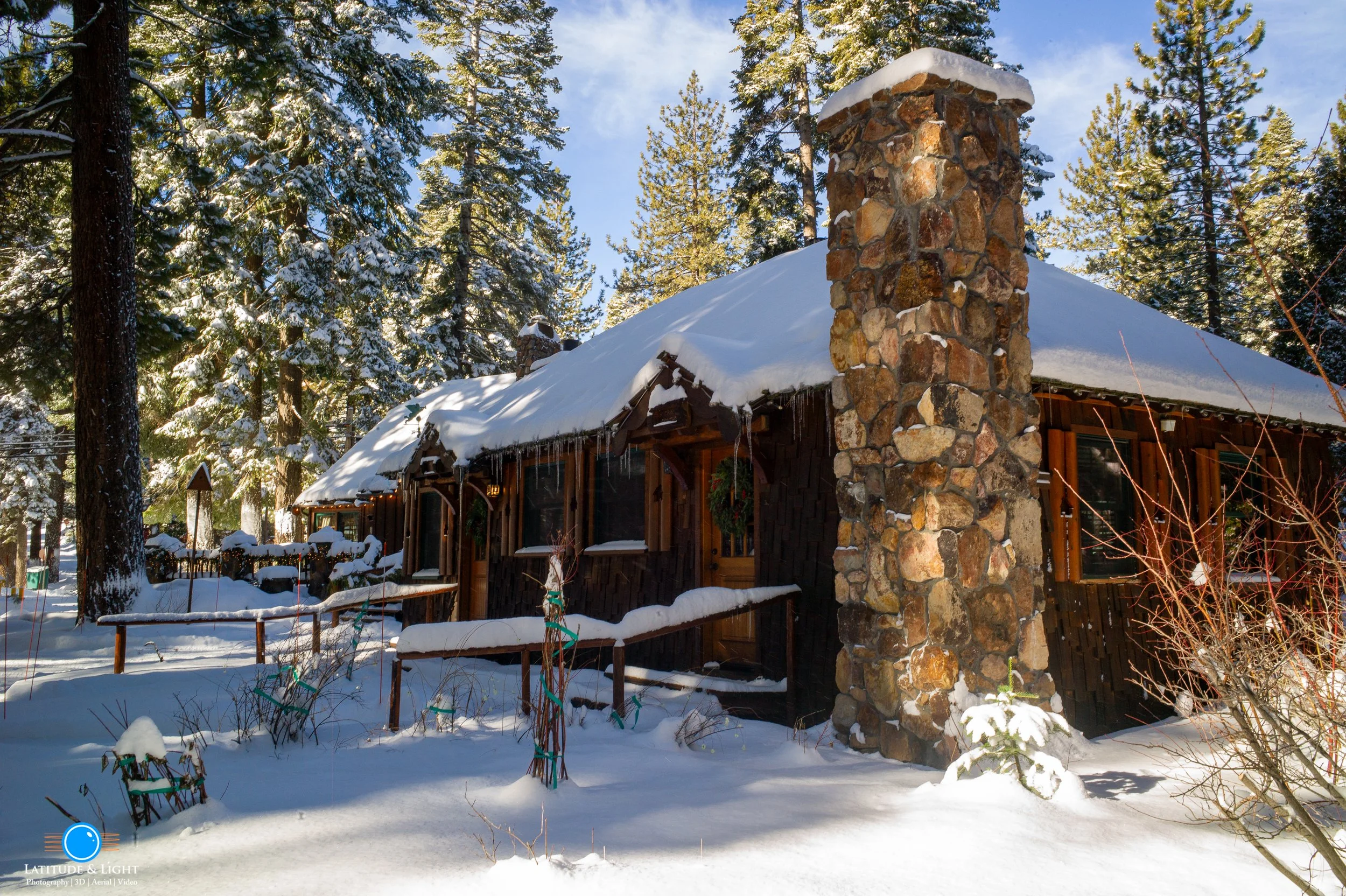 A cozy snow-covered cabin in a forest with tall pine trees in Tahoe City. The cabin has a stone chimney and a wooden railing, with snow accumulating on the roof and surrounding ground.