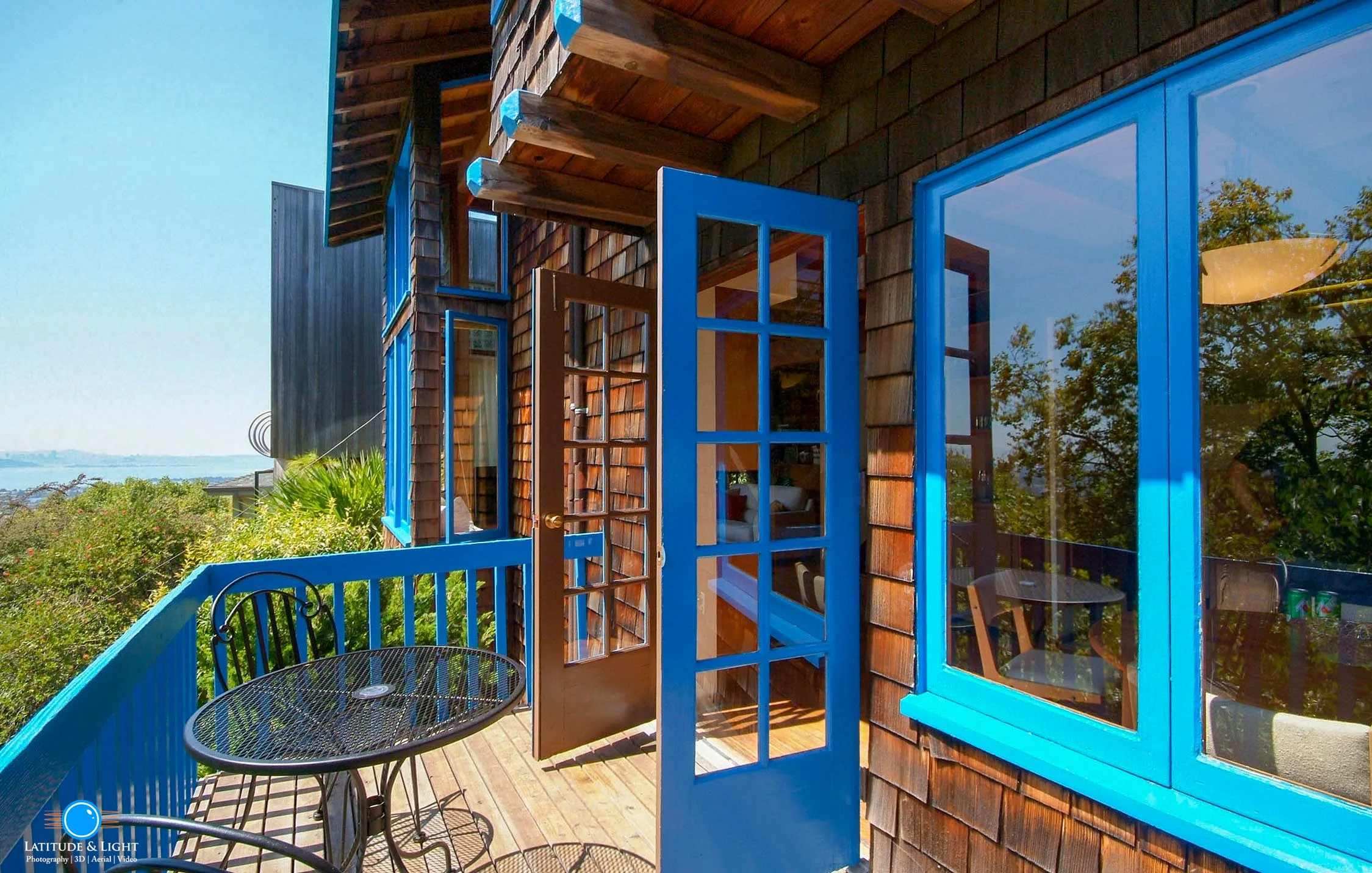 Pacific coast balcony with a black metal table and chair, blue window frames, and a wooden door, overlooking greenery and water in the distance.