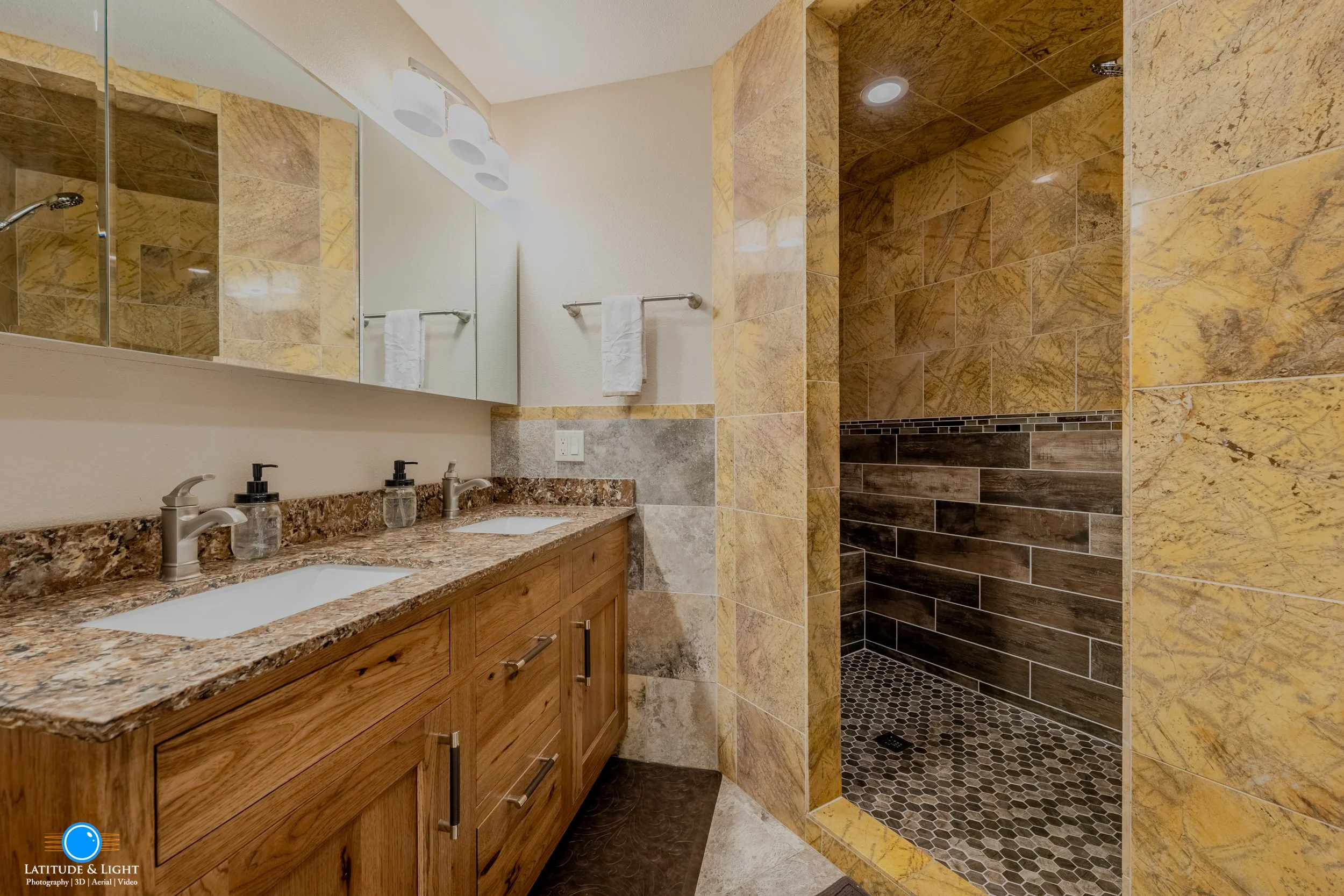 Bathroom with a double sink vanity, granite countertop, and a walk-in shower with brown and yellow tile walls and hexagonal floor tiles.