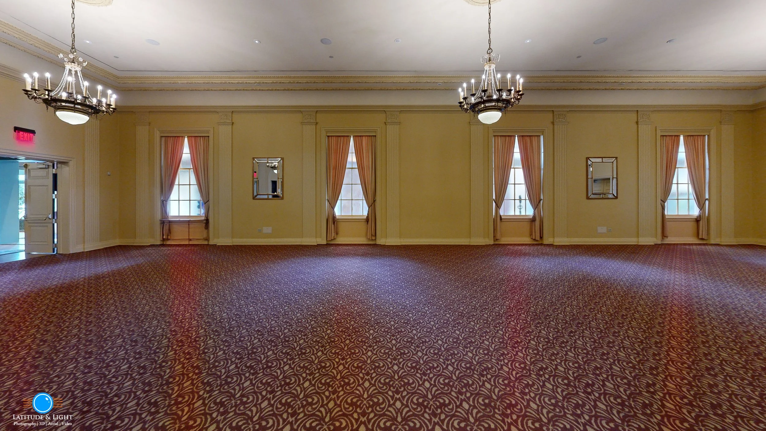 Empty ballroom in Walla Walla with beige walls, multiple windows with pink curtains, chandeliers, and patterned red carpet.