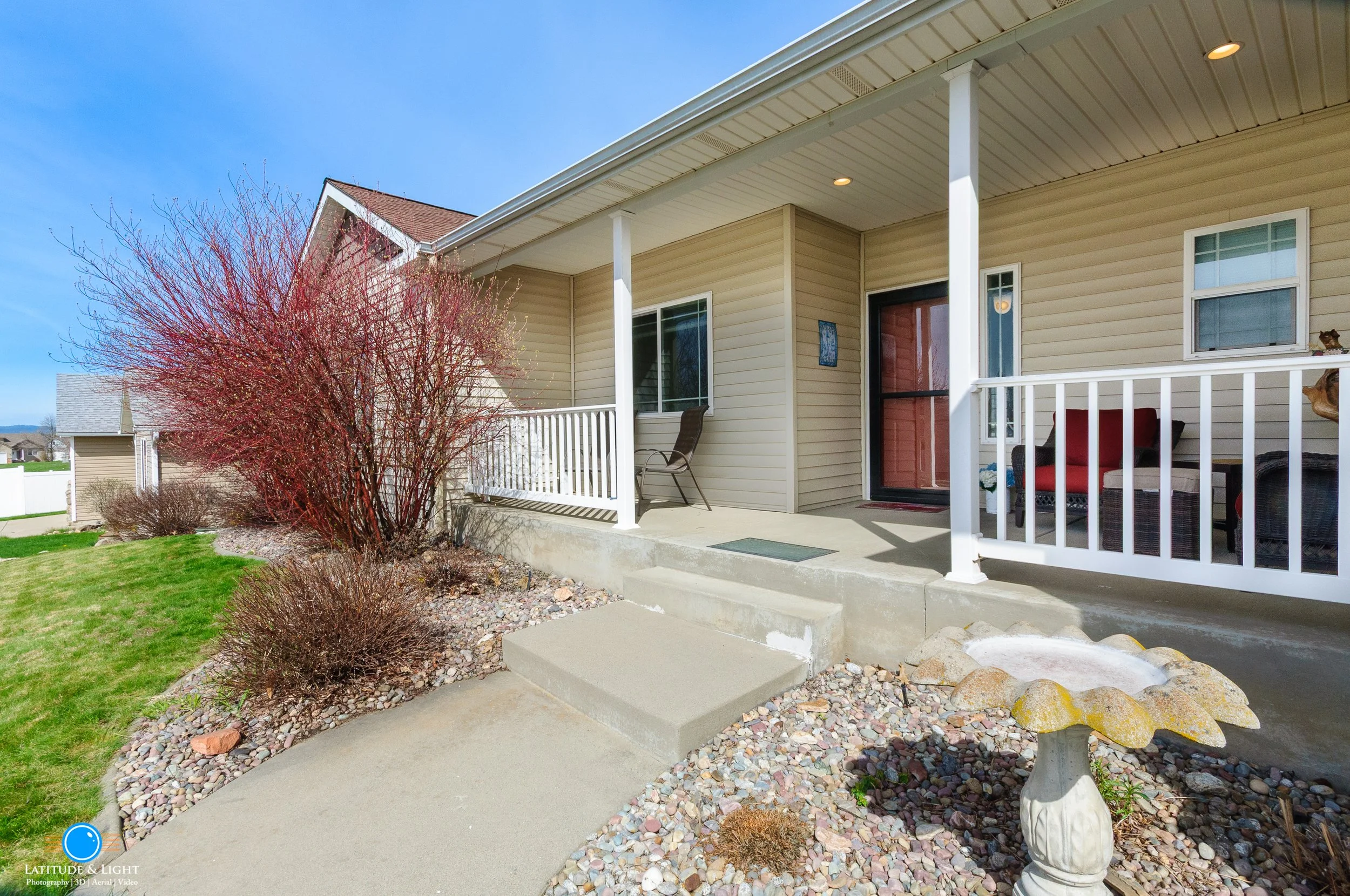 Front porch of a Post Falls house with white railing, a red door, two chairs, and a small birdbath in a landscaped yard with bushes and a tree, under a blue sky.