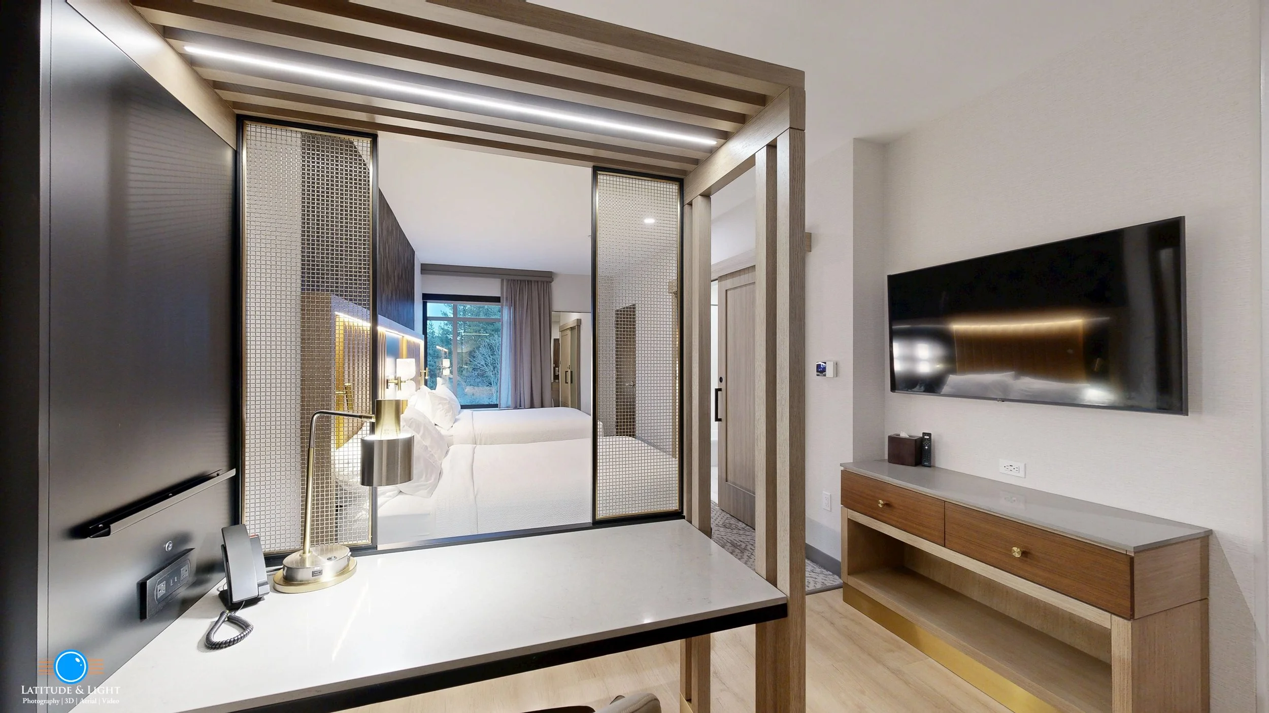 A modern hotel room with two beds, a large flat-screen TV on the wall, and a workspace with a lamp and phone. The beds are separated by a divider with a gold accent light, and there are large windows with curtains in the background.