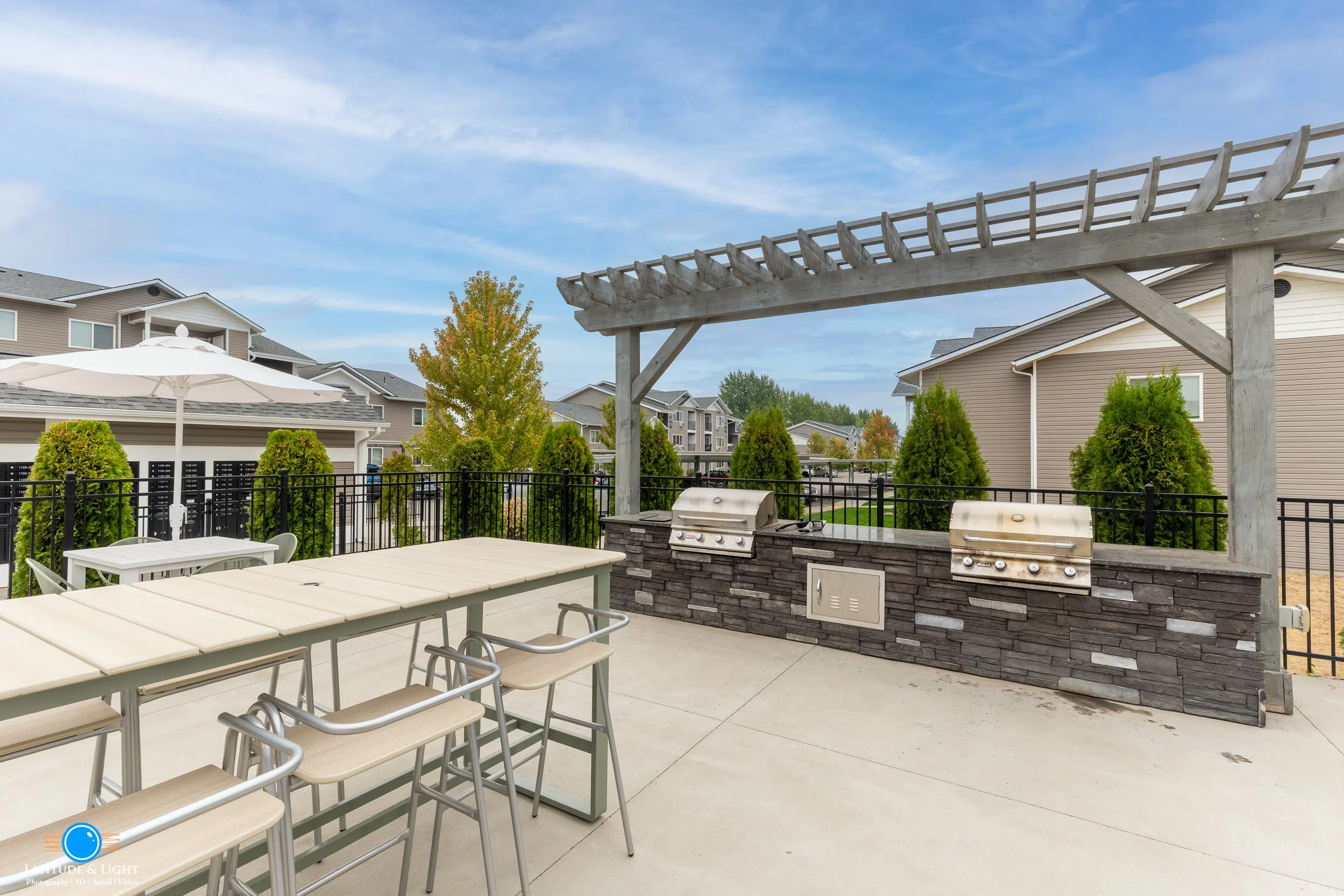 Outdoor patio with a grilling station, a table with chairs, and a canopy frame against a backdrop of suburban houses and green trees.