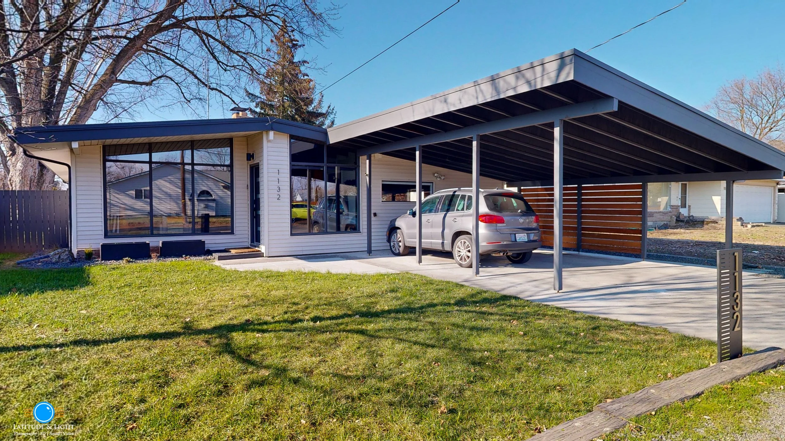 A modern single-story house with a carport, gray siding, large windows, and a small front yard with green grass and a mailbox displaying house number 1132.