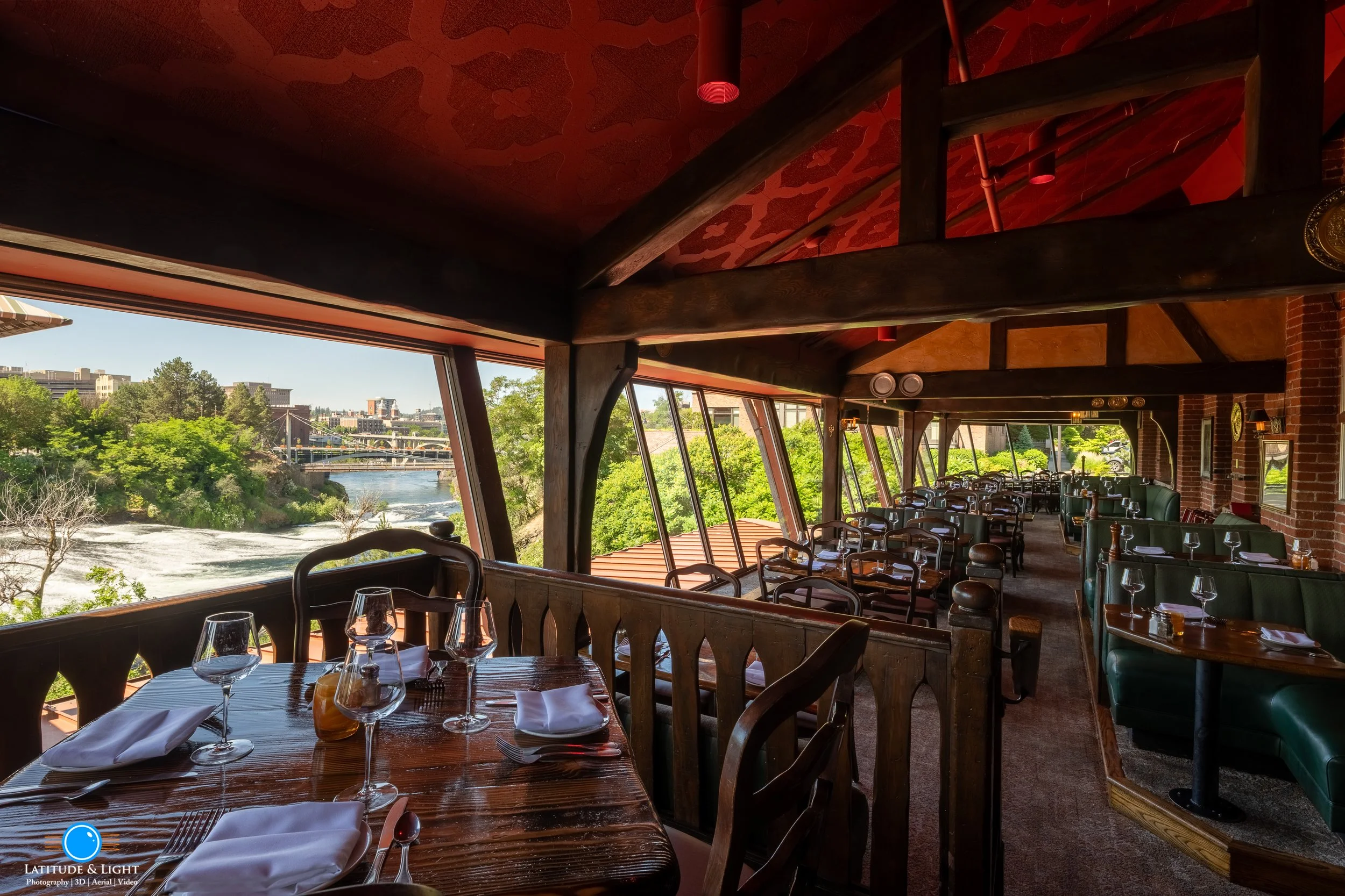 Interior of a Spokane restaurant with a large window showing a river, trees, and a bridge outside. The dining area has wooden tables set with wine glasses, napkins, and utensils, along with green booths and wooden chairs.