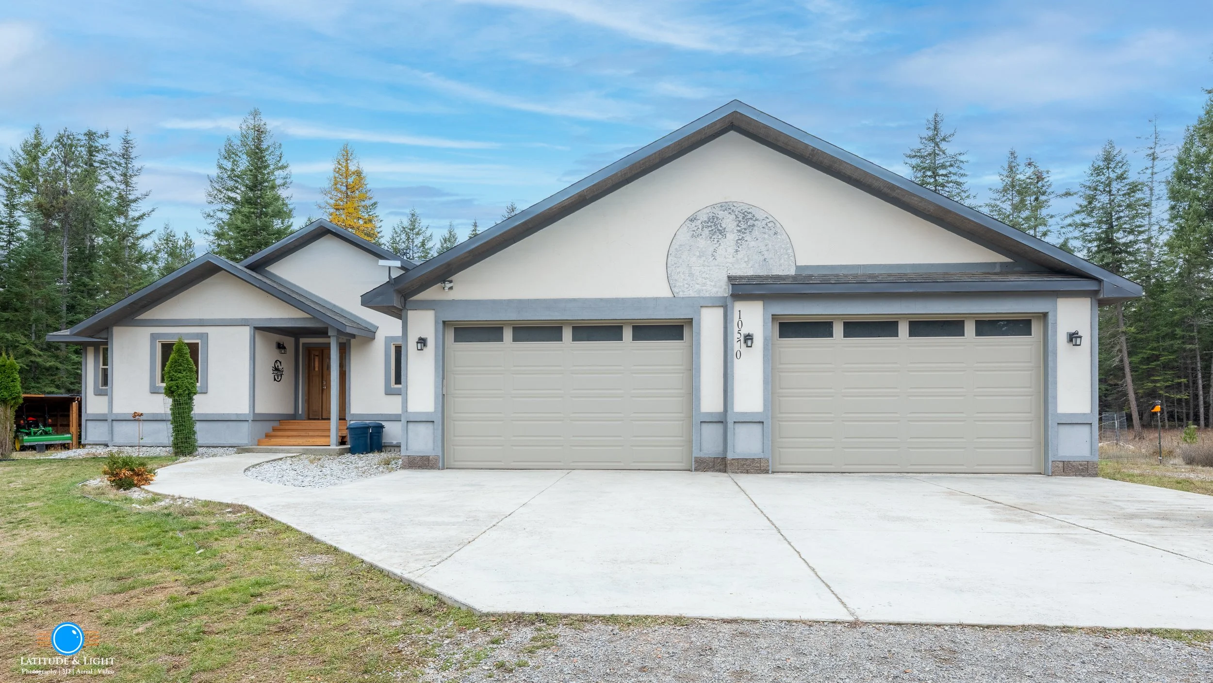 Front view of a modern house near Coeur D'Alene with a three-car garage, white exterior walls with gray trim, and a wooden front door. The house has a landscaped front yard with a concrete driveway and green grass, surrounded by trees.