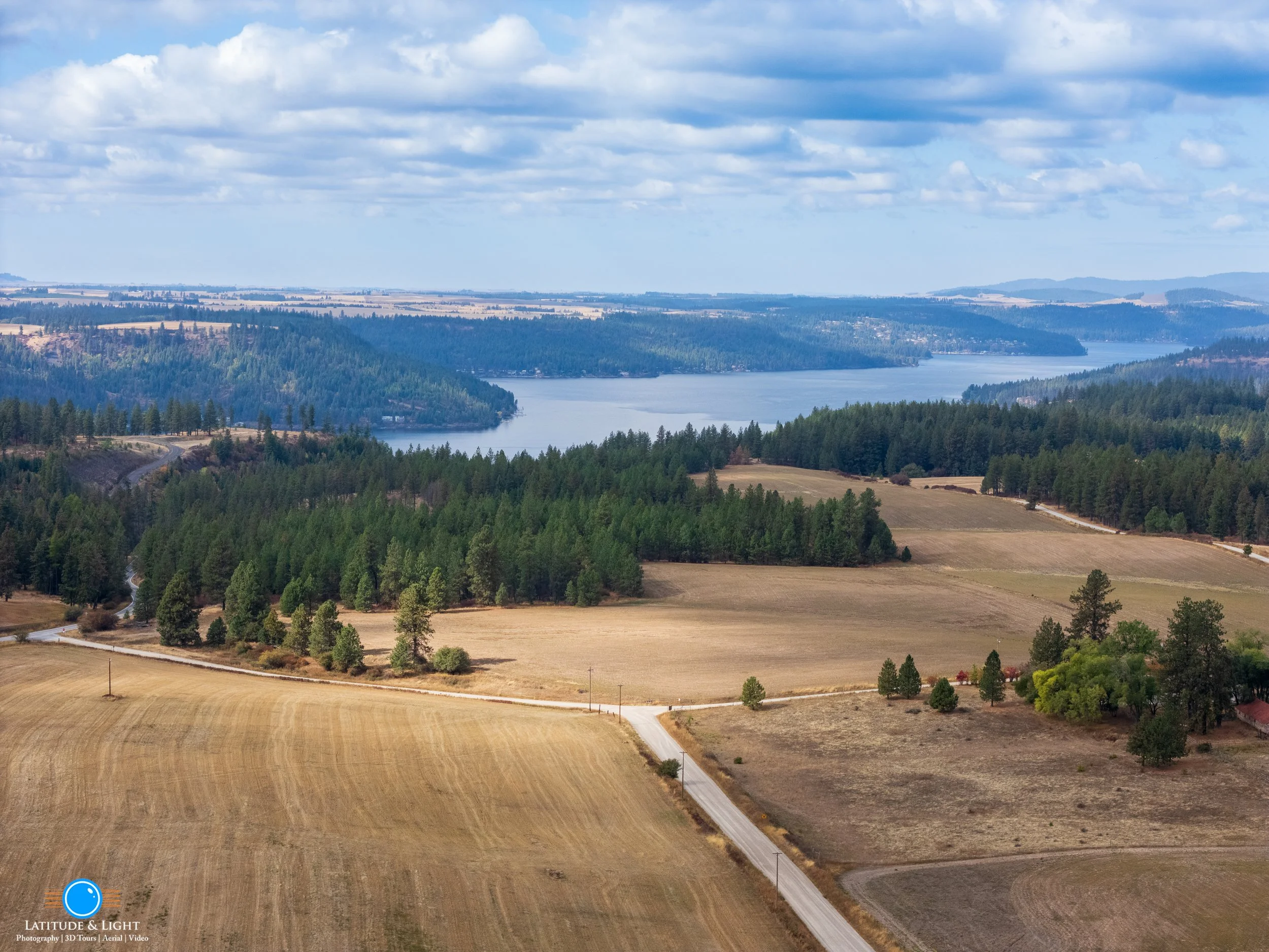 A wide landscape view of southern Lake Coeur D'Alene surrounded by hills and forests, with fields and roads in the foreground under partly cloudy skies.