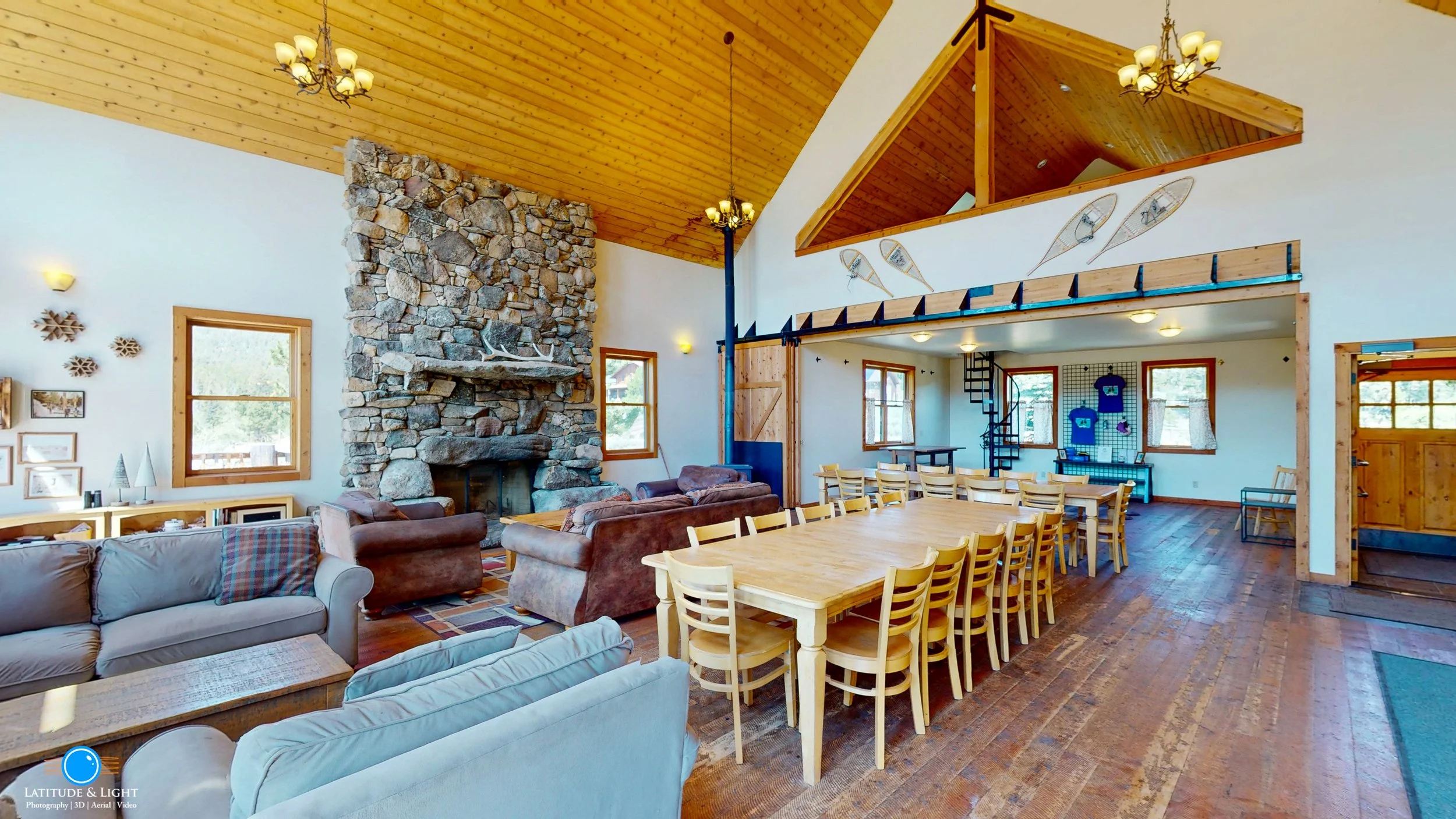 Interior of a rustic lodge with a large stone fireplace, wooden ceiling, and multiple seating areas. Wooden dining tables and chairs are arranged for dining, and snowshoes are mounted on the wall. Large windows let in natural light.