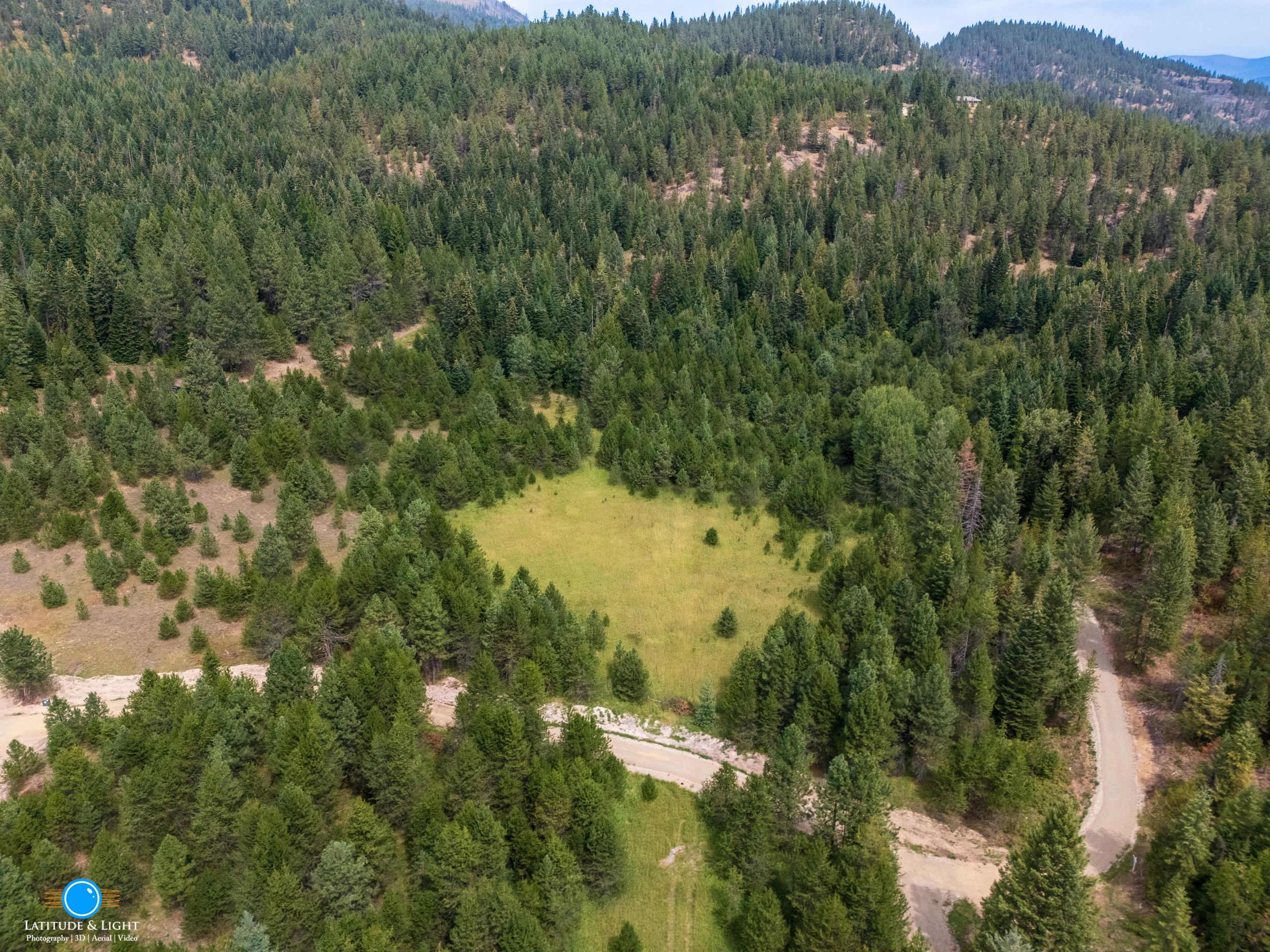 An aerial view of a forested mountain landscape with a dirt road running through the trees and ending in a clearing with grass.