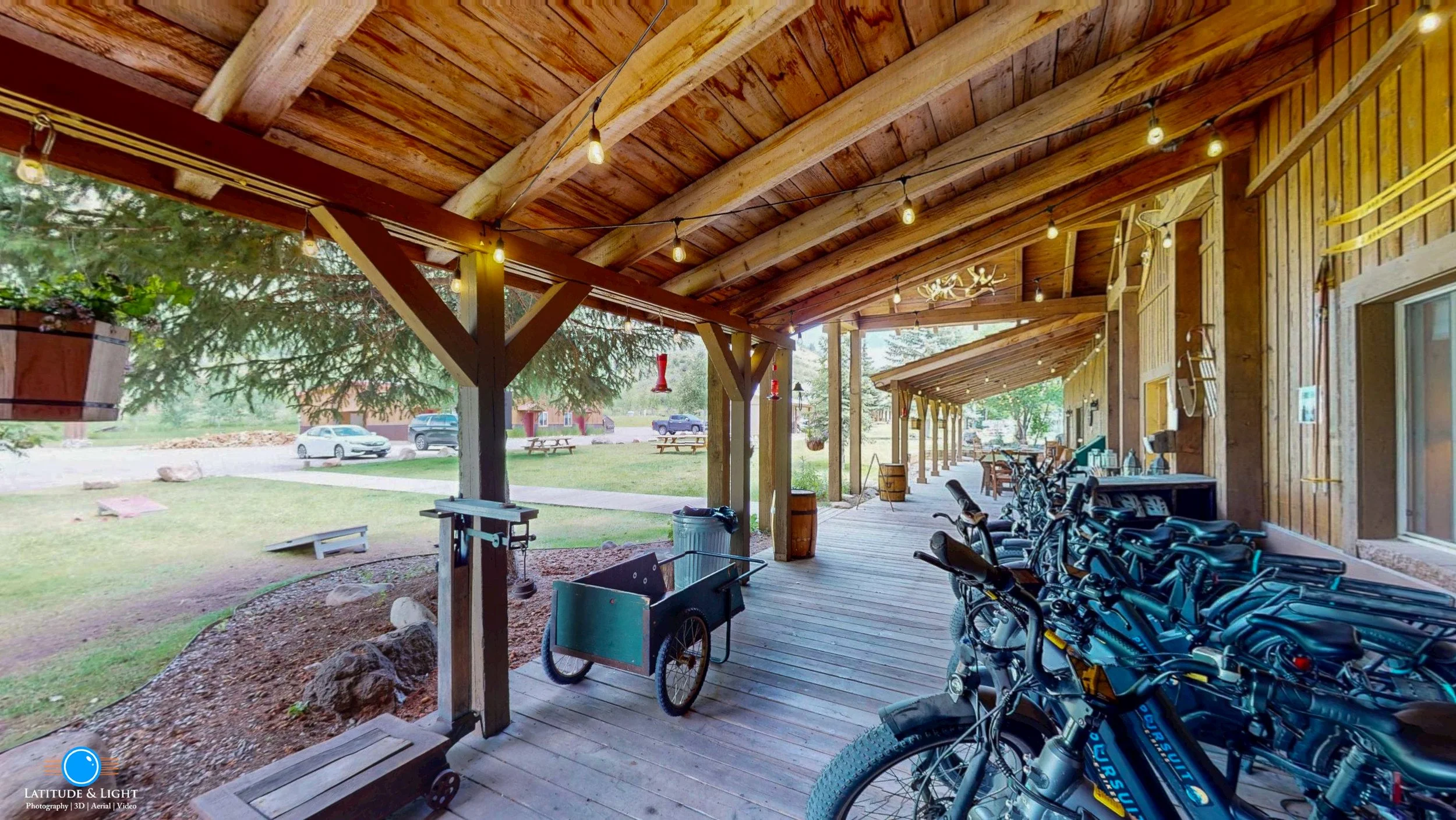 Covered outdoor patio area with wooden ceiling, string lights, bicycle rack with bikes, wooden tables and chairs, and a grassy area with parked cars in the background.
